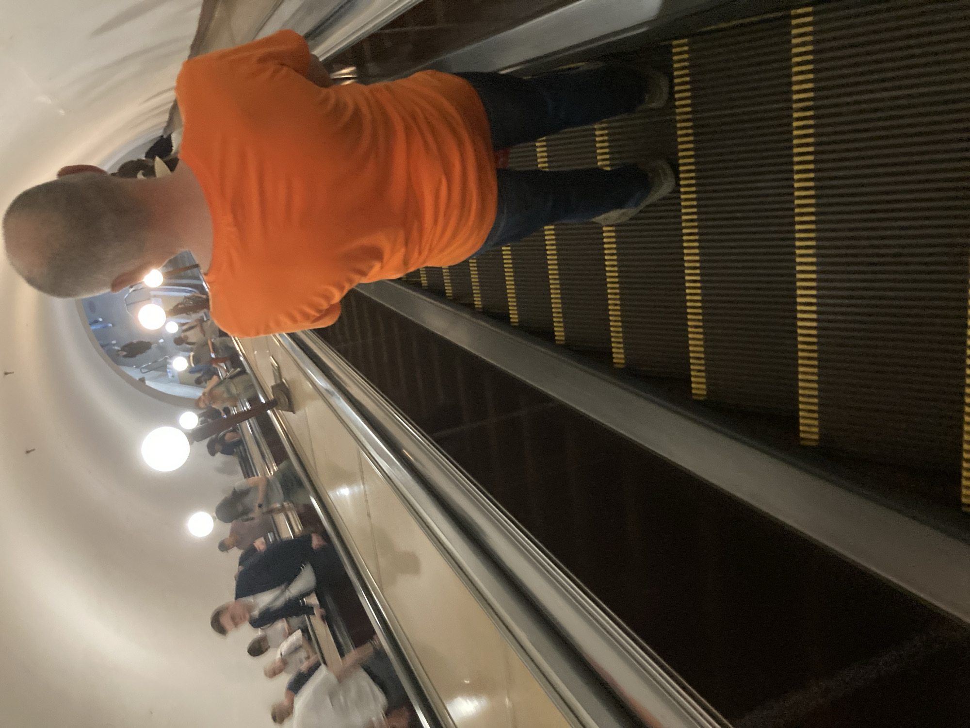A man with a shaved head is riding an escalator in a Moscow Metro station. He wears an orange shirt, dark blue jeans, and light-colored shoes. The escalator has black treads with yellow stripes and silver railings. Other people are visible on the adjacent escalator. The station has a curved white ceiling with spherical lights.