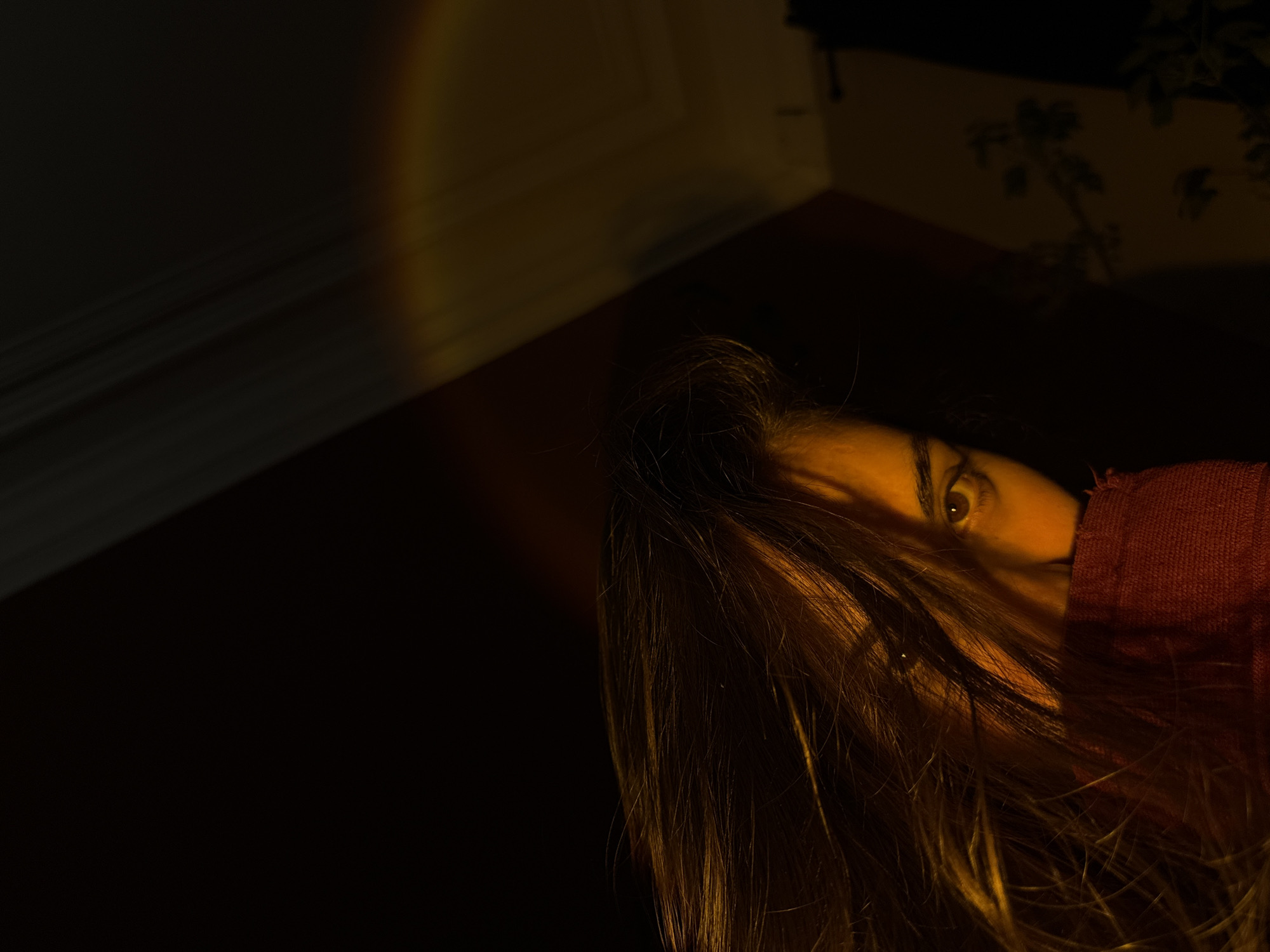 A dimly lit interior scene presents a partial view of a person with long, brown hair obscuring much of their face, but two eyes are visible and focused on the viewer. The person is partially covered by a red fabric garment. A diffuse, yellow light source illuminates the top left corner of the image and part of the person's hair. The background includes portions of walls with what appears to be floral wallpaper in a dark corner. There is no visible indication that the location is Moscow, Russia.