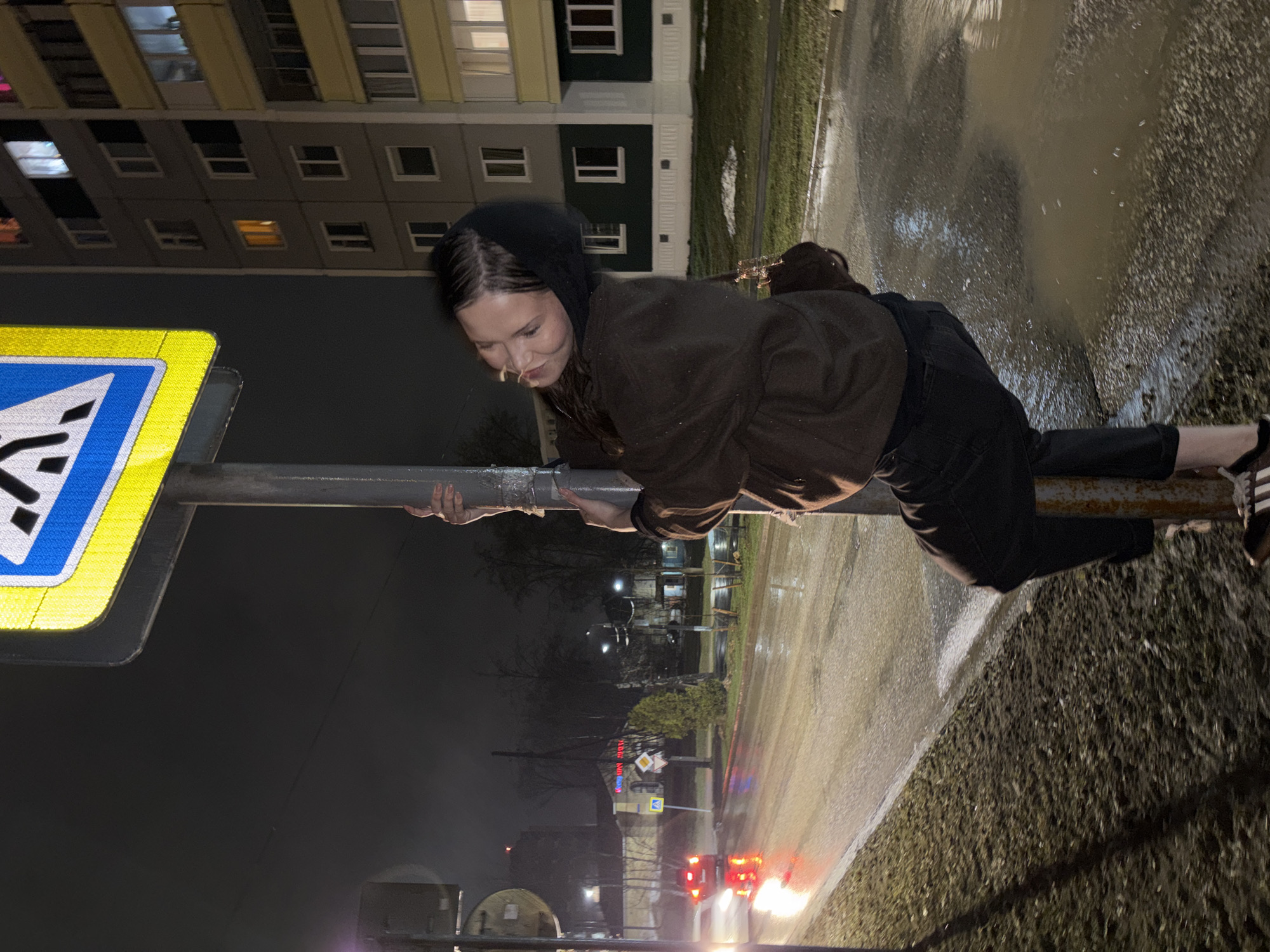 A young woman in a dark hoodie and brown jacket is lying across a pedestrian crossing signpost at night in Perm, Russia. She's wearing dark pants and Adidas sneakers. Her face is turned toward the sign, and she appears to be holding onto the post with both hands. A building with lighted windows stands in the background, and a wet road reflects the lights of passing vehicles. The scene is illuminated by streetlights and vehicle headlights. The ground appears wet.