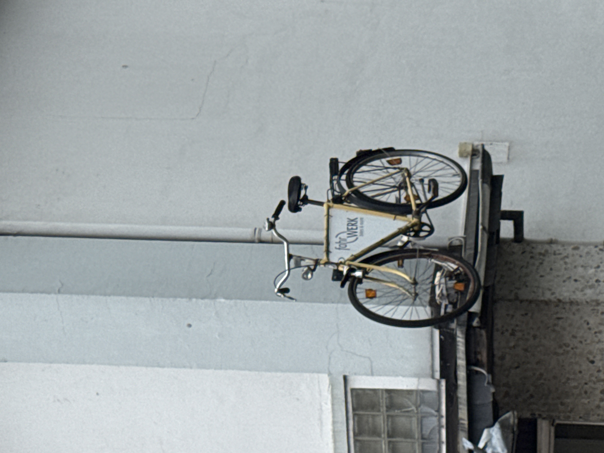 A yellow bicycle is mounted on the exterior wall of a building in Bassum, Germany. The bicycle is positioned horizontally, with its front wheel pointing left and its rear wheel pointing right. A sign that reads "fahrWERK" is attached to the frame of the bicycle. The building appears to be made of concrete, and there is a window with multiple panes visible to the left of the bicycle. A downspout runs vertically along the wall behind the bicycle. The scene is set against a light gray wall.