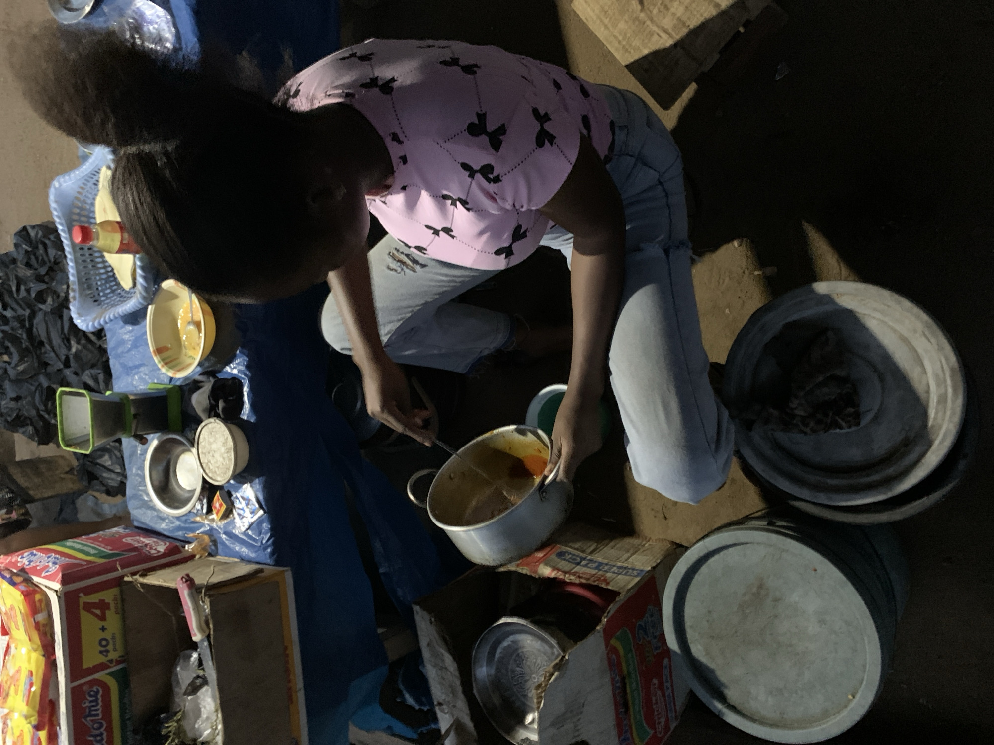 The image captures a person cooking or preparing food in a dimly lit, informal setting, likely at night. The individual, with dark hair pulled back, is wearing a pink short-sleeved top with black bow patterns and light blue ripped jeans. They are squatting or kneeling, actively stirring a reddish-orange substance in a metal pot with a spoon, holding the pot with their left hand.
The ground appears to be dirt or an unpaved surface, covered in parts by a blue tarp. Surrounding the person are various objects:
To their left, a light blue plastic basket holds a bottle with a red cap and a yellow bowl. Nearby, a pile of dark fabric or bags and a small green funnel-like object are visible.
Several metal bowls and pots are scattered around, some empty, others containing unknown contents.
On the right side of the frame, two large, gray lidded containers or drums are present.
Two cardboard cartons are used as makeshift surfaces. One is a prominent red and yellow carton for "Indomie" instant noodles, with "40+4 pacha" and "SUPER PACK" visible on its sides.
The overall scene suggests a functional food preparation area, possibly a street food stall or a home kitchen setup in a less formal environment. The low lighting, strong shadows, and types of containers indicate a nighttime setting. The visible "Indomie" packaging is common in Nigeria, aligning with the provided location context of Kabba, Nigeria.