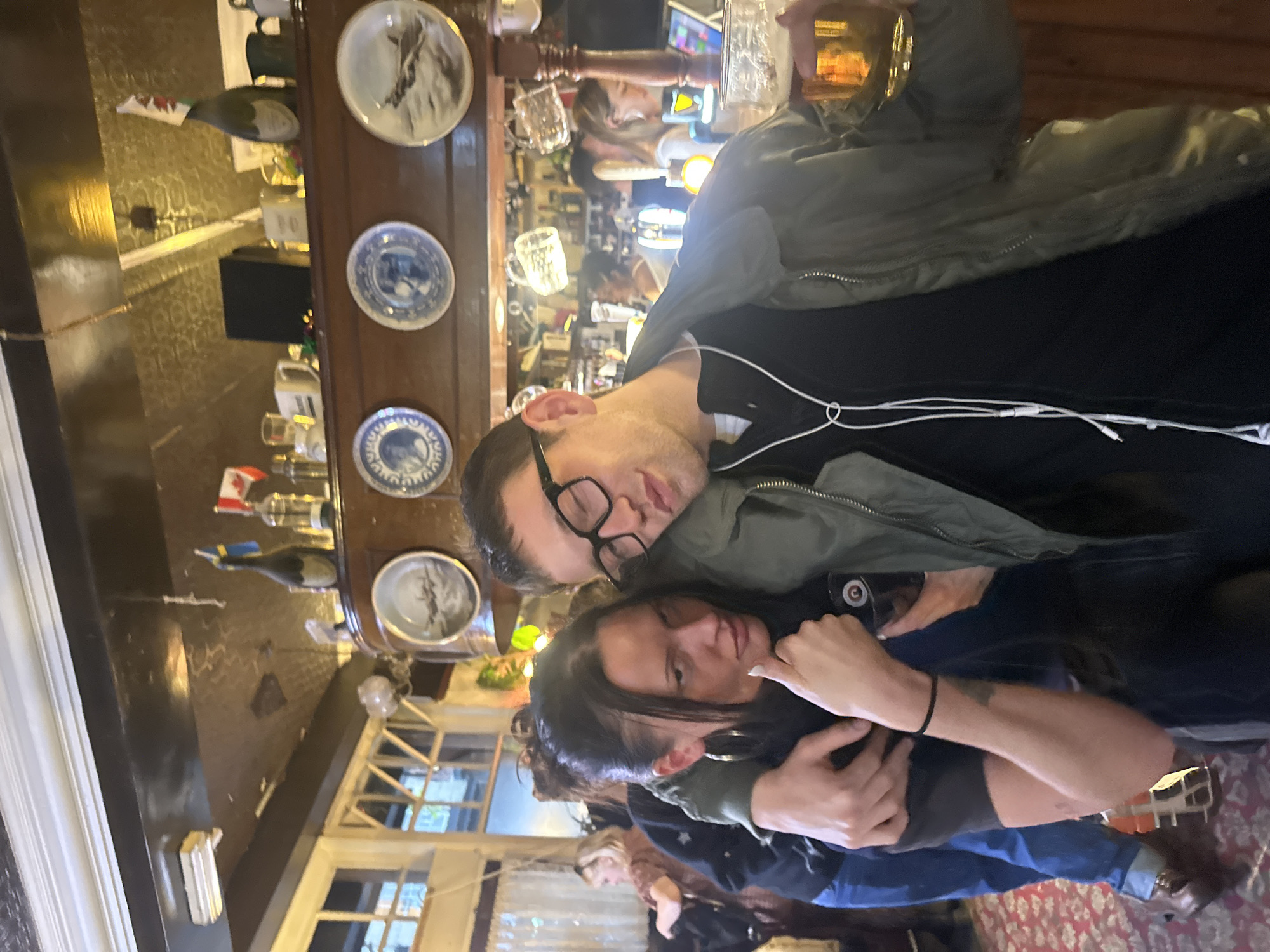 The image shows a man and a woman embracing in what appears to be a traditional British pub. The man is wearing glasses, a black shirt, and a grey jacket. He is holding a glass of beer. The woman is wearing large hoop earrings and a black jacket. Behind them is a bar with various glasses, bottles, and decorative plates displayed. A window is visible in the background, suggesting a view to the outside. The scene suggests a social gathering or outing in a pub.
