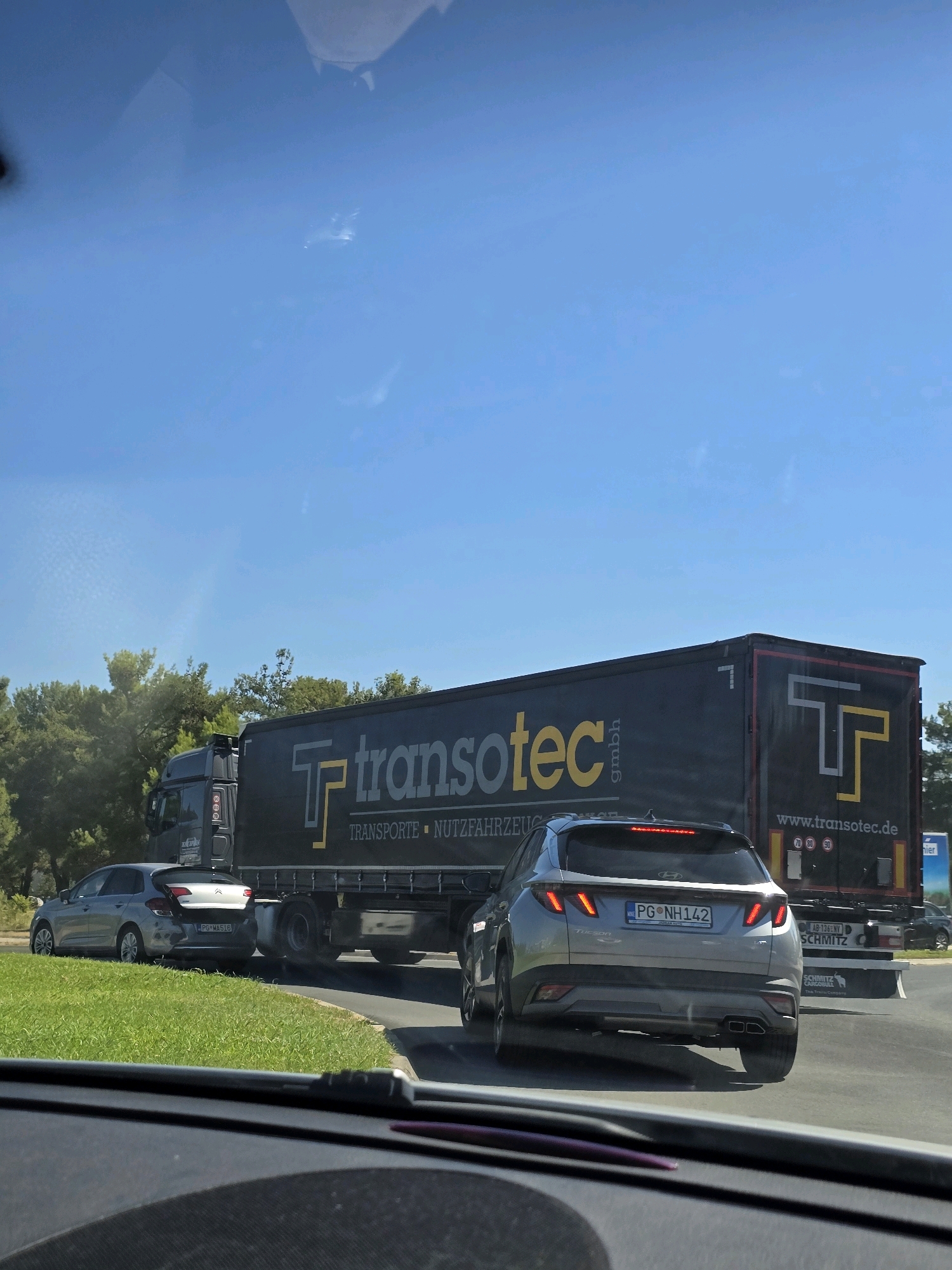 The image shows a sunny day with a clear blue sky. In the foreground, a vehicle's dashboard is visible. Ahead, a silver Hyundai Tucson is driving on a road, its brake lights illuminated. To the left of the Tucson, a silver Citroën C4 is also on the road. A large, black semi-trailer truck with "transotec" branding is to the right of these cars. The truck has a website address "www.transotec.de" and speed limit signs (70, 80, 90) on its rear. The scene appears to be a highway or a main road, with green grassy areas and trees lining the sides. There are no people clearly visible in the image. The license plate of the Hyundai Tucson reads "PG NH 142". The license plate of the Citroën C4 reads "PG MA516". Given the presence of the license plates starting with "PG", which is the standard prefix for Podgorica, Montenegro, and the context of European road signs and vehicle makes, it is plausible this scene is in or around the city of Podgorica.