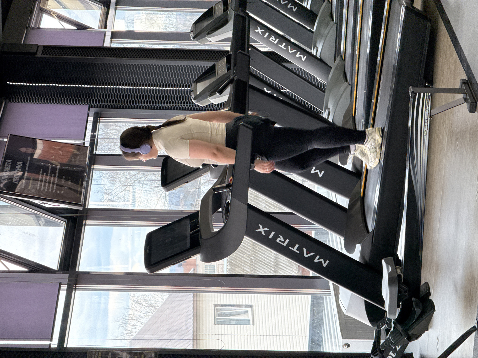 A woman with headphones is using a Matrix brand treadmill in a gym with large windows. The gym is located in Votkinsk, Russia, based on the view of the buildings outside the windows and the artwork visible. Other treadmills are visible in the background. The woman is wearing dark athletic leggings and a light-colored shirt.