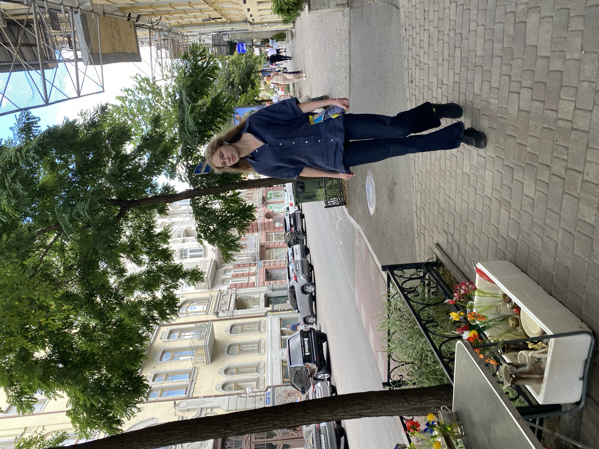 A young woman with blonde hair and glasses walks down a brick sidewalk in a city. She is wearing a dark blue short-sleeved shirt and dark blue pants. She is holding a blue and yellow object in her right hand. In the background, cars are parked along the street, and buildings with architectural details line the opposite side of the street. Trees with green foliage are present along the sidewalk. To the left of the woman, a table with flowers in vases and a white container with various items is visible. The lighting suggests it is daytime.