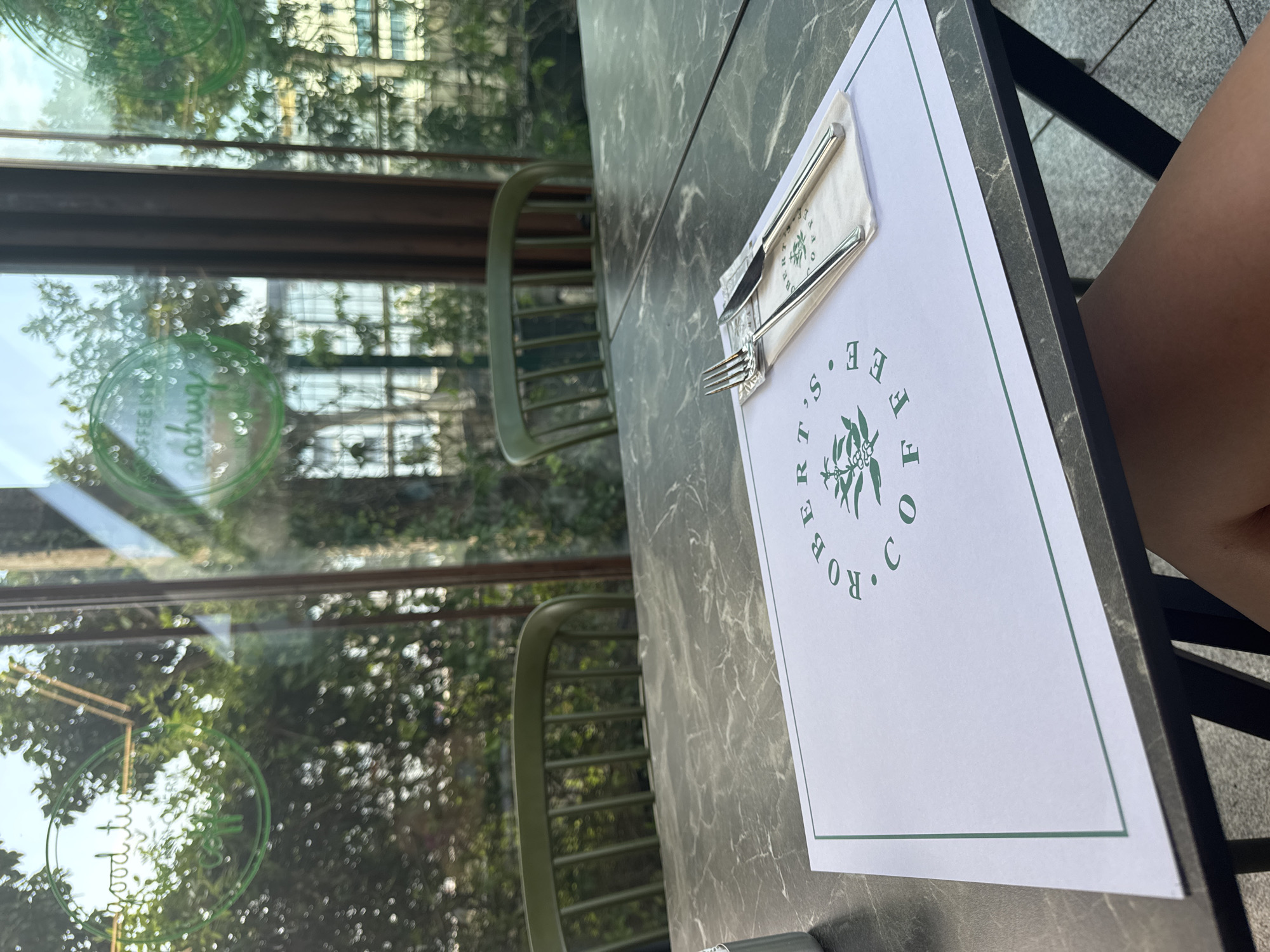 The image shows a table setting at a cafe, likely "Robert's Coffee" based on the placemat. The table has a dark, marble-patterned surface. A white placemat with a green border is laid out, featuring the "Robert's Coffee" logo in green text and a stylized illustration of coffee plants. A fork and knife, wrapped in a white napkin, are placed on the placemat. In the background, through a large window, greenery and trees are visible, suggesting an outdoor or garden-like setting. Two empty green chairs are also visible through the window. The overall impression is of a relaxed cafe environment, possibly in Antalya, Turkey, given the brand's presence there. The lighting suggests it is daytime, with sunlight filtering through the leaves.