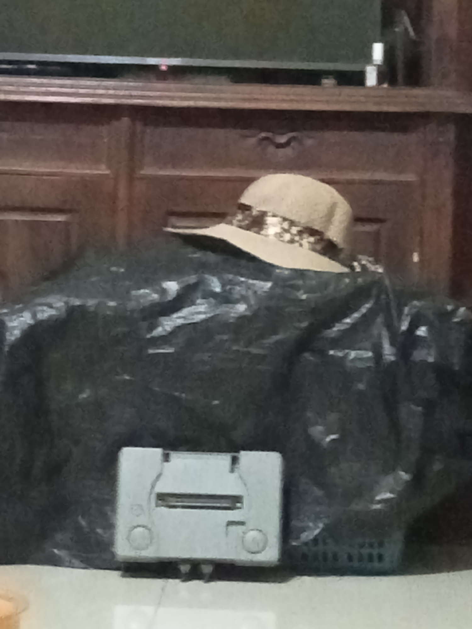 A light-colored straw hat with a patterned band sits atop a pile of black plastic sheeting. In front of the sheeting, a grey video game console is visible. The scene is set against a dark wooden cabinet with a dark television screen visible above it. The overall impression is one of an indoor storage area, possibly in a residential setting. The exact location in Grabag, Indonesia cannot be determined from the image.