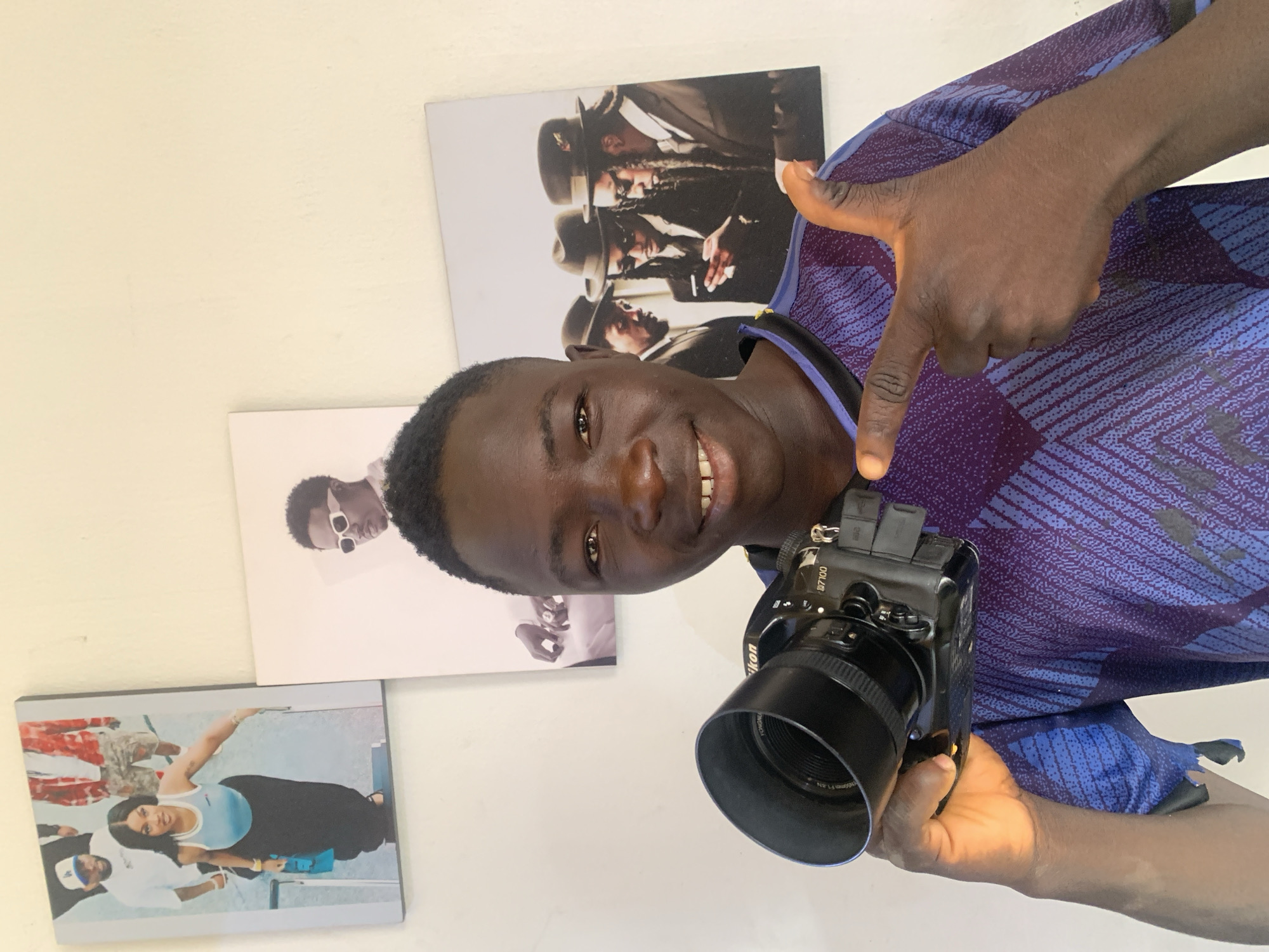 A young man, likely in Jos, Nigeria, is captured in a close-up, chest-up portrait, smiling and looking directly at the viewer. He has dark skin, short dark hair, and a warm, inviting smile. He wears a blue and purple patterned short-sleeved shirt. His right hand is raised, with his index finger pointing forward, possibly gesturing towards the camera he holds or the act of photography itself.
In his left hand, he holds a black Nikon DSLR camera. Visible text on the camera clearly identifies it as a "Nikon D7100" body, equipped with a "Nikon AF-S NIKKOR 50mm 1:1.8G" lens. A small accessory is mounted on the camera's hot shoe.
The man is positioned indoors against a plain, light cream-colored wall. Behind him, three framed pictures or posters are displayed. To his upper right, a vertical poster shows three men, possibly a musical group, wearing hats and dark clothing, with dreadlocks. One appears to be smoking. To his mid-left, another vertical poster features a man with dark skin, wearing sunglasses. Below that, to his lower left, a horizontal poster depicts a group of people outdoors, possibly near a pool or beach, with a smiling woman in a light blue top and dark bottoms as the central figure, and another man in a white cap and t-shirt visible. The overall scene suggests a creative or personal space, highlighting the man's engagement with photography.