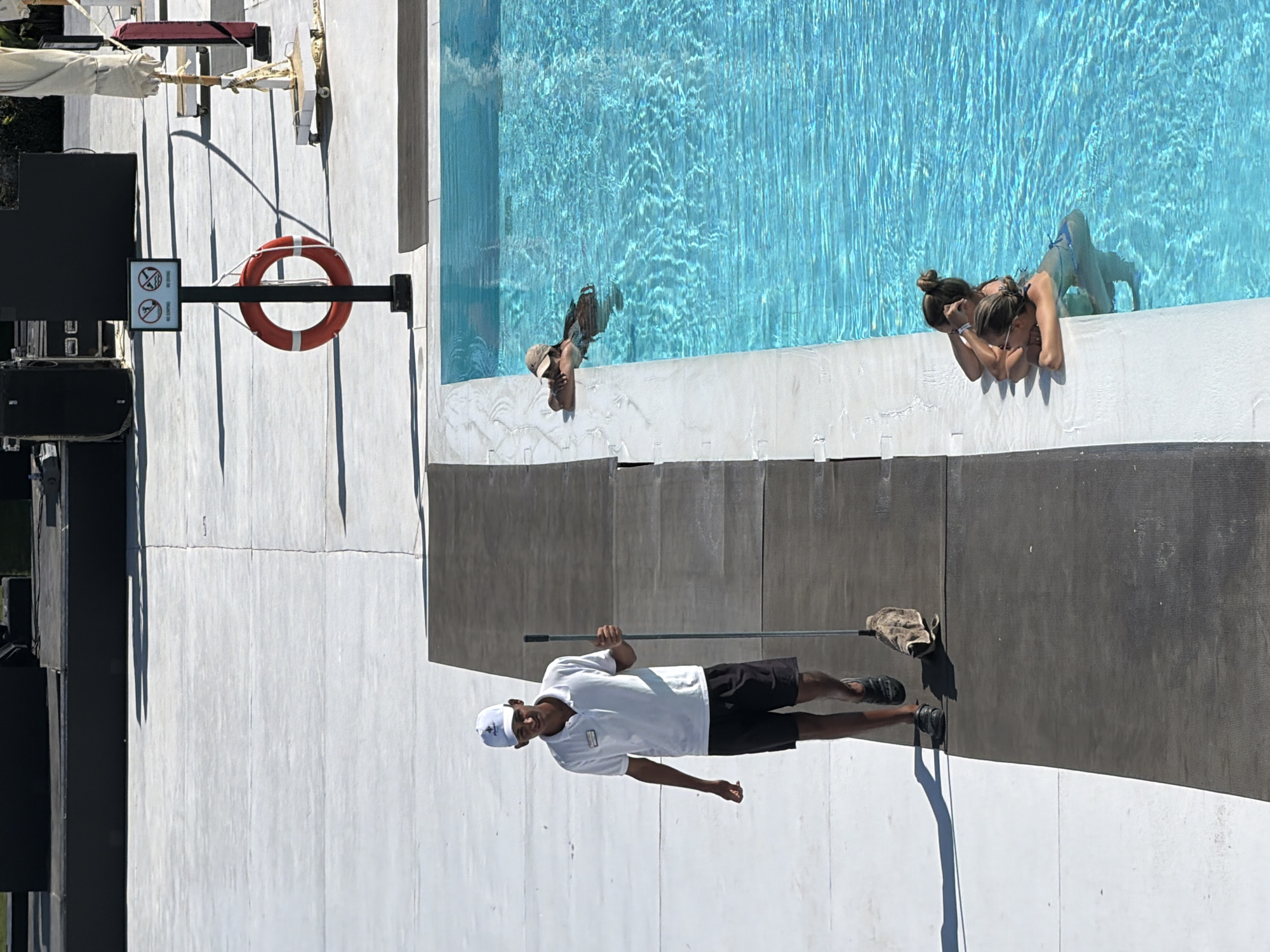 This image captures a sunlit outdoor scene at a resort in Sharm El Sheikh, Egypt. In the foreground, a young man wearing a white polo shirt, black shorts, and a white baseball cap is walking and pushing a mop. He appears to be an employee, possibly a lifeguard or groundskeeper, as he is carrying a cleaning tool. To his right, a vibrant blue swimming pool is visible, with several women relaxing in the water near the edge. One woman, wearing a cap, leans on the pool edge. Further back in the pool, two other women are visible. The poolside area is paved with white tiles and dark gray sections, suggesting a modern resort design. In the background, there are large black speaker cabinets, a life preserver ring hanging on a post with a "no jumping" and "no diving" sign, and partially visible white umbrellas. The overall impression is of a leisure area in a warm, possibly tropical, climate.
