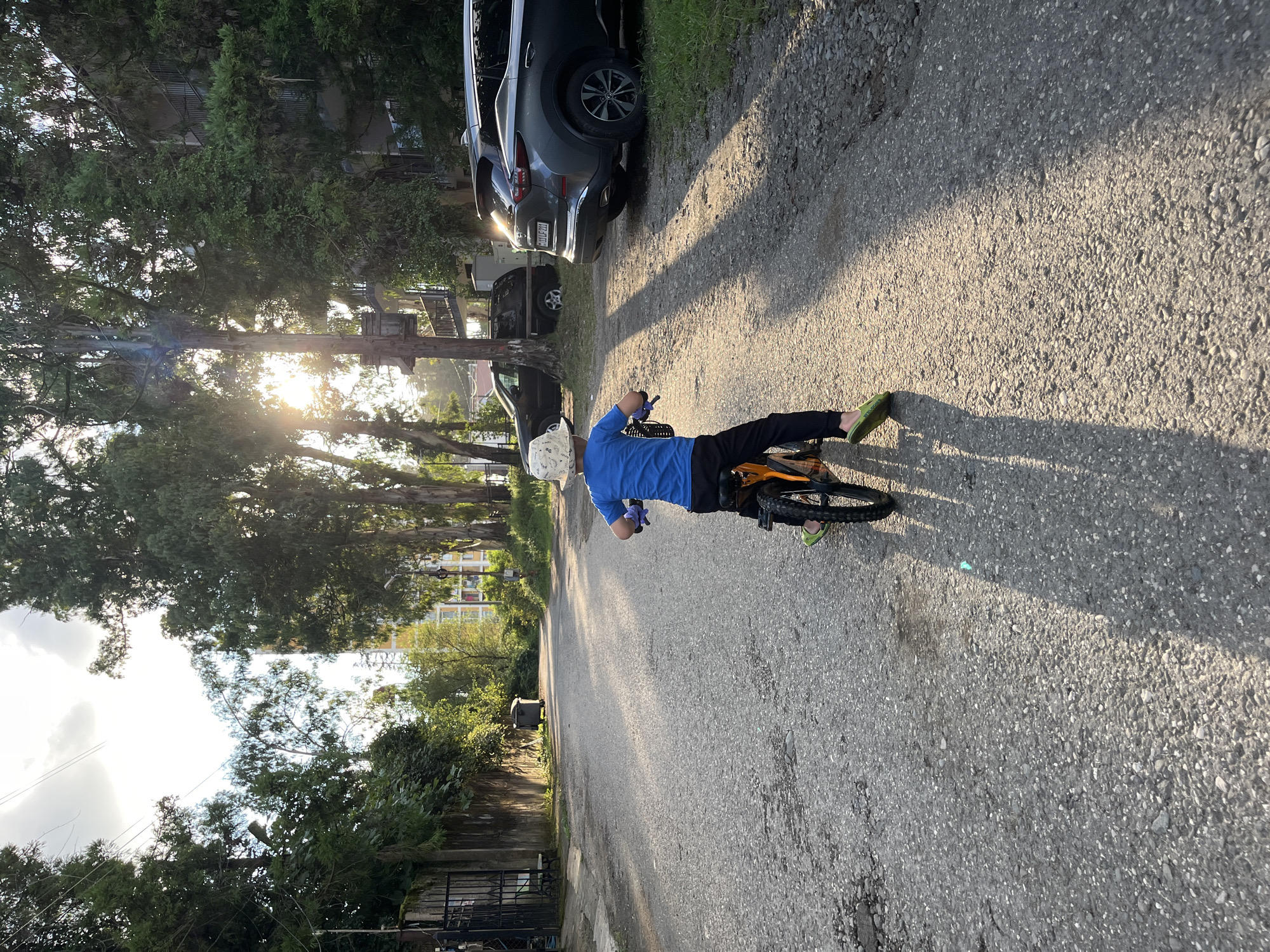 A young boy wearing a white patterned sun hat, a blue t-shirt, and black pants rides an orange bicycle down a paved road in Ureki, Georgia. The boy is facing away from the camera, with his hands on the handlebars and his feet on the pedals. He wears green slip-on shoes. Tall trees with lush foliage line the road, casting long shadows across the asphalt. Sunlight filters through the trees, creating a dappled effect. On the right side of the road, two cars are parked: a dark SUV in the foreground and a lighter colored SUV behind it. In the background, buildings and more trees are visible. The sky is bright, with some clouds. The scene appears to be a quiet residential street during the late afternoon.