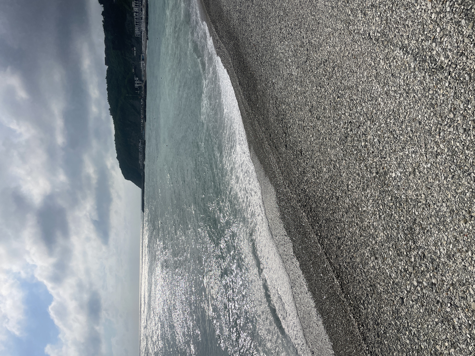 The image shows a beach scene in Olginka, Russia. The beach is composed of small, grey pebbles. The water is a pale, reflective color with small waves lapping at the shore. In the background, a heavily vegetated cliff rises, with buildings visible on top. The sky is overcast with grey clouds, though a patch of blue sky is visible in the lower left corner. The photograph has a slight vertical orientation.