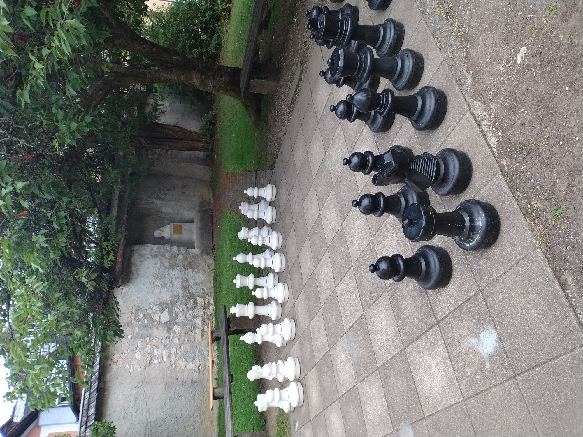 An outdoor oversized chess set is arranged on a tiled patio in Hallstatt, Austria. The chess pieces are predominantly white and black, with the white pieces positioned on the left side of the patio and the black pieces on the right. A stone wall, partially covered in ivy, forms the background, with a wooden bench in front of it. A large tree with dense foliage partially obscures a building with a wooden shingled roof. The ground around the patio is a mix of grass and dirt. No people are present in the image.