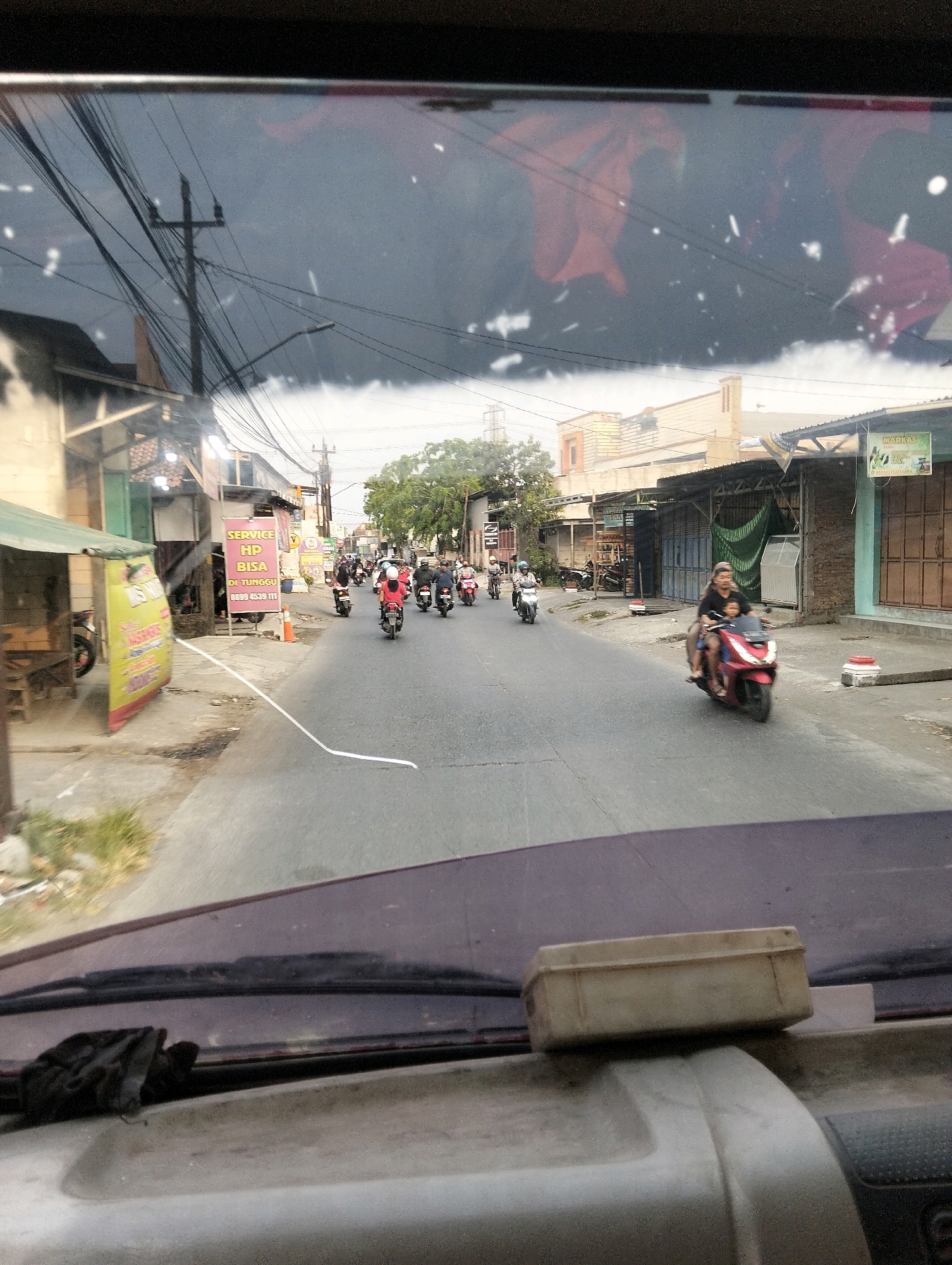 The image depicts a street scene in Semarang, Indonesia, likely captured from inside a vehicle. The foreground shows the dashboard of the vehicle, with a dirty windshield. A small, light-colored rectangular object, possibly a container or box, rests on the dashboard.
The middle ground of the image shows a street with several people riding motorcycles. On the right side of the street, a person is riding a red motorcycle with a child as a passenger. The child appears to be wearing a red shirt. Further down the road, other motorcyclists are visible, some of whom appear to be wearing helmets.
The background reveals a row of buildings on both sides of the street. On the left, there are several small shops or kiosks, one with a green awning and a yellow banner advertising services. Another shop has a pink banner with Indonesian text. On the right, there are buildings with closed storefronts and awnings. Power lines crisscross above the street, and a utility pole is visible on the left. Trees are present in the distance, suggesting an urban or suburban environment. The sky appears overcast, contributing to a muted lighting condition. The overall impression is of a bustling street in Indonesia.