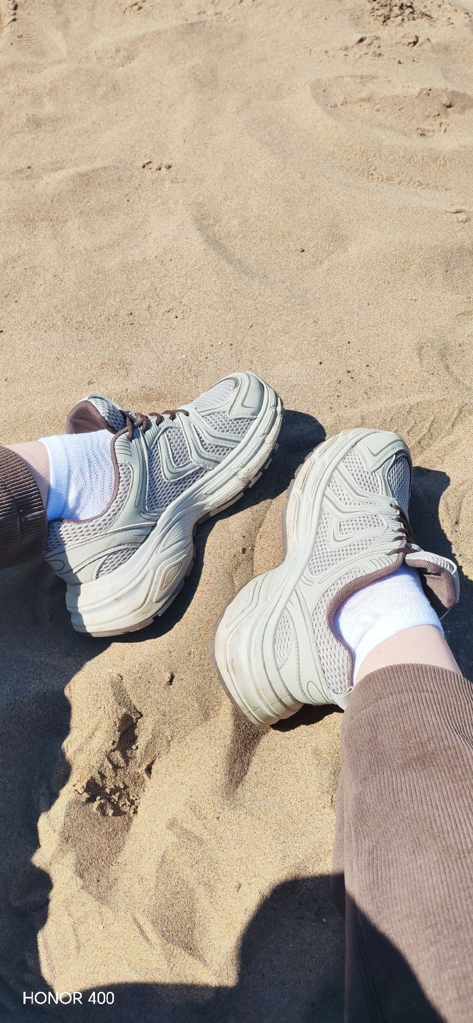 The image shows a pair of light grey athletic shoes with white socks, worn by a person sitting on a sandy surface, presumably a beach. The person is wearing brown corduroy pants. The sand is light tan and there are shadows indicating sunlight. The lower left corner of the image has the text "HONOR 400". The exact location within Noviy, Russia is not discernible from the image.