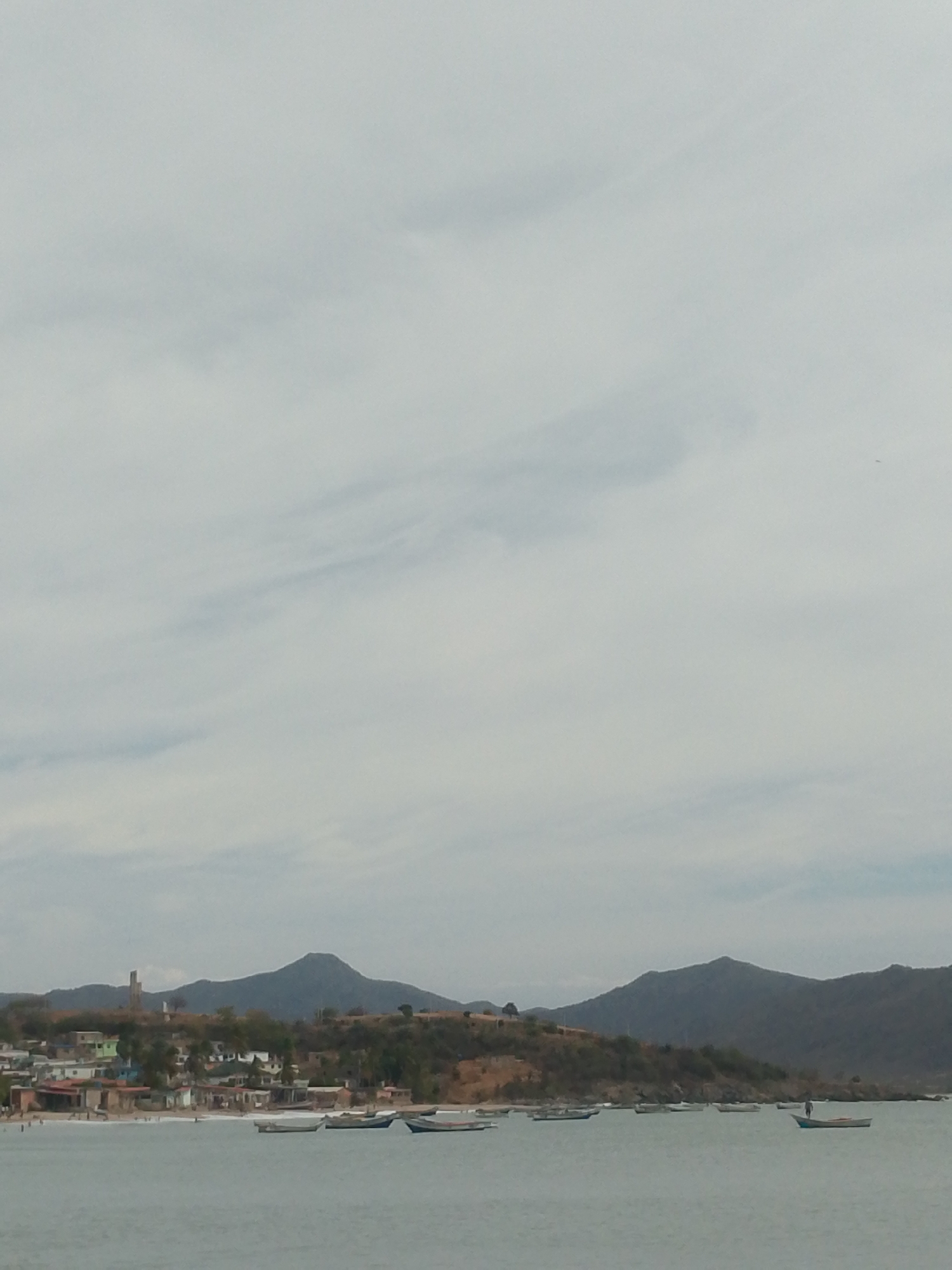 The image shows a coastal scene in Juan Griego, Venezuela. In the foreground is the ocean, with several small boats anchored. One boat has a person standing in it. Further back is a coastline with a beach, small buildings, and houses. A prominent tower-like structure is visible on the hillside among the buildings. In the background, there are hills. The sky is overcast and pale.