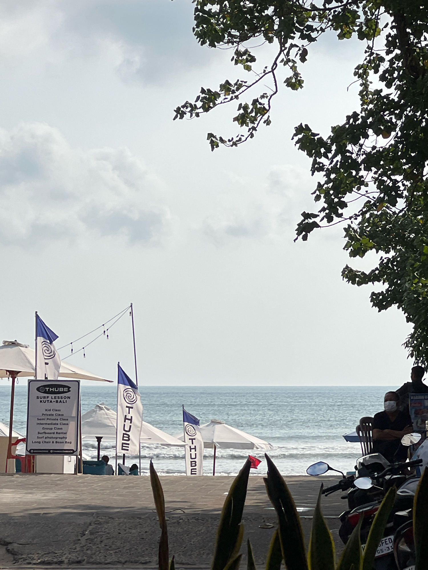 This image shows a beach scene in Kuta, Indonesia. The foreground features a paved walkway and a patch of greenery, with several motorcycles parked to the right. Beyond the walkway, beach umbrellas and flags are visible, indicating a business offering surf lessons. A sign displays "THUBE SURF LESSON KUTA BALI" and lists various services. The ocean stretches to the horizon under a cloudy sky, with gentle waves lapping the shore. Several people are present in the mid-ground, some sitting under umbrellas and one wearing a face mask. Trees with green foliage frame the upper right portion of the scene.