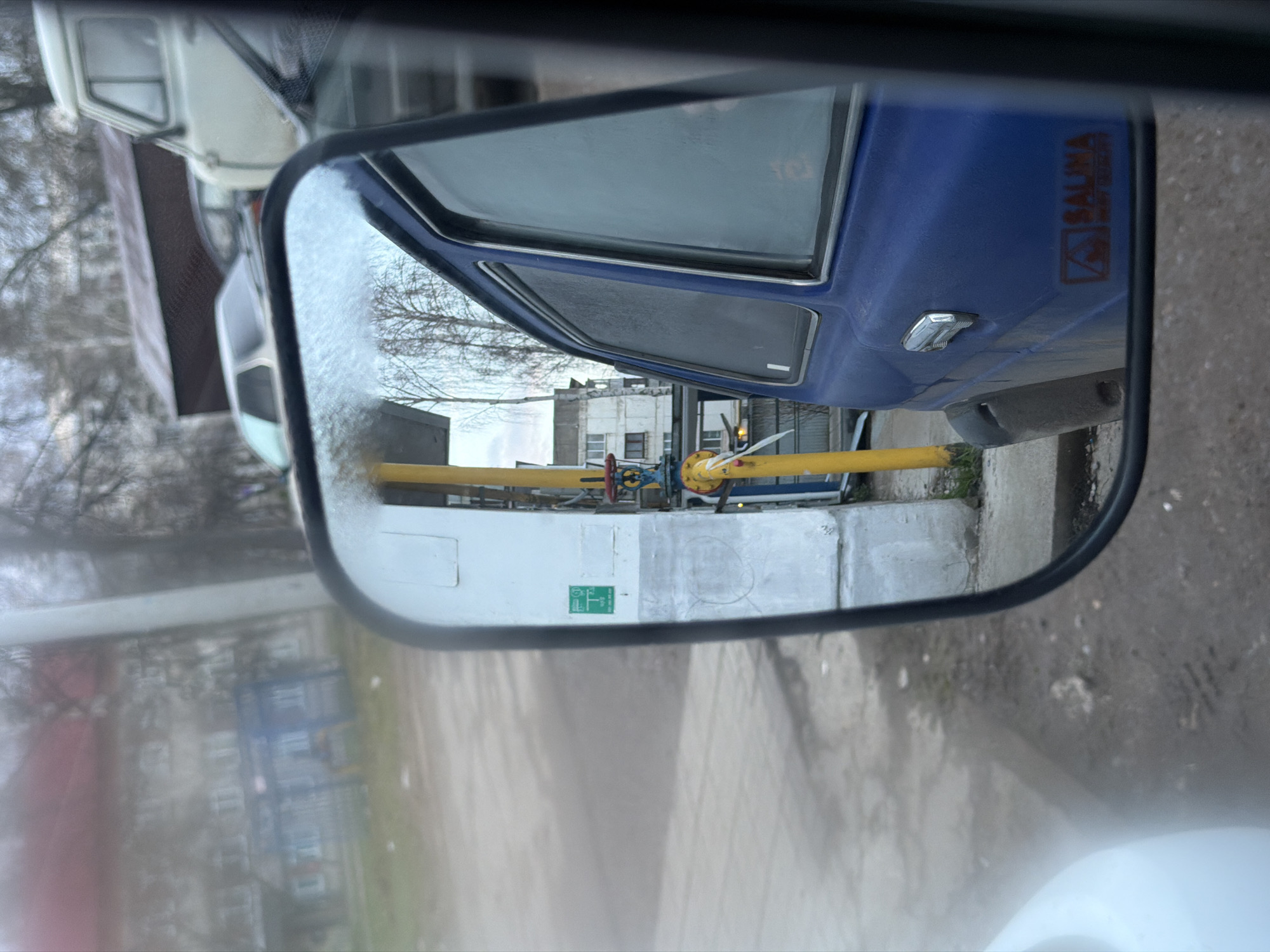 The image, taken from inside a vehicle via the side mirror, depicts a view of the street and nearby structures in what appears to be the city of Chaikovsky, Russia.
In the side mirror's reflection, a blue vehicle with the word "SALINA" visible is seen. Next to the blue car is a white structure with yellow gas pipes running along it. A building with visible windows is in the background.
Outside the mirror, the interior of the vehicle from which the photo was taken is visible, including parts of the dashboard and the passenger seat. The exterior view outside the mirror is blurred. The ground seems to be made of stone or concrete, showing signs of wear. The sky appears cloudy or overcast.
The photo lacks any clear sign of people or immediate human activity. The setting is an urban environment with a street, buildings, and infrastructure components like gas pipelines.