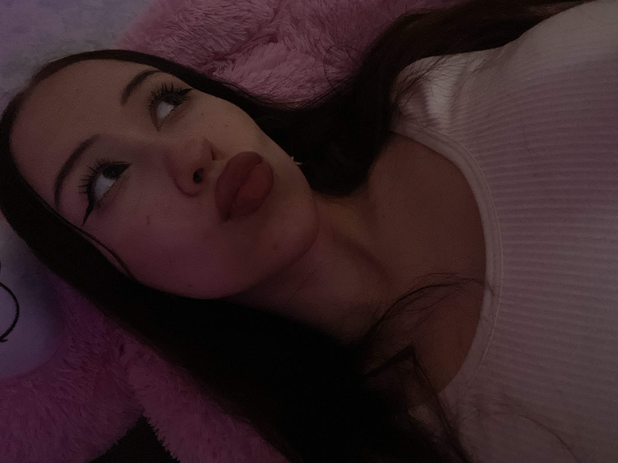 A young woman with long, dark hair and prominent eyeliner is captured in a close-up, over-the-shoulder selfie. She is wearing a white ribbed tank top. Her lips are puckered, and her gaze is directed off to her right. The background is a soft, fuzzy pink texture, likely a blanket or plush toy. The lighting is dim, with a pinkish hue casting over the scene. There is no indication of the city of Votkinsk, Russia, or any specific location.