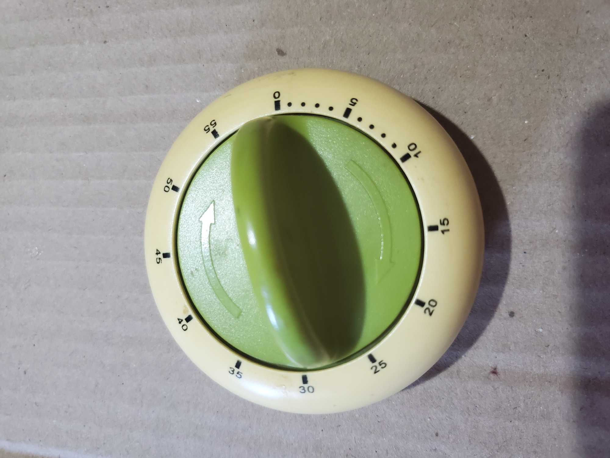 The image shows a close-up, overhead view of a kitchen timer. The timer is round and cream-colored with a green dial in the center. The dial has an arrow pointing towards the '0' mark, indicating it is set to zero. The outer rim of the timer is marked with numbers from 0 to 55, indicating minutes. There are small black tick marks between the numbers, representing individual minutes. The timer is resting on a textured, brown surface, which appears to be cardboard or a similar material. There are no people or other objects visible in the frame. The lighting suggests an indoor setting. The image does not provide any information about its location in Chișinău, Moldova.