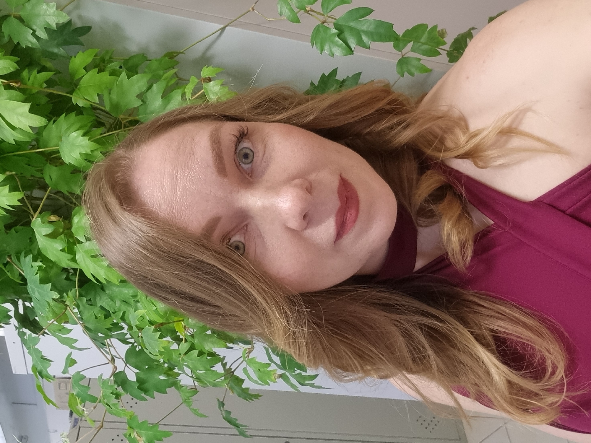A woman with light skin and long, light brown hair poses in front of a wall covered in green leafy vines. She is wearing a maroon dress. The background includes a white wall with what appears to be a cabinet below. The location is not depicted or verifiable in this image.