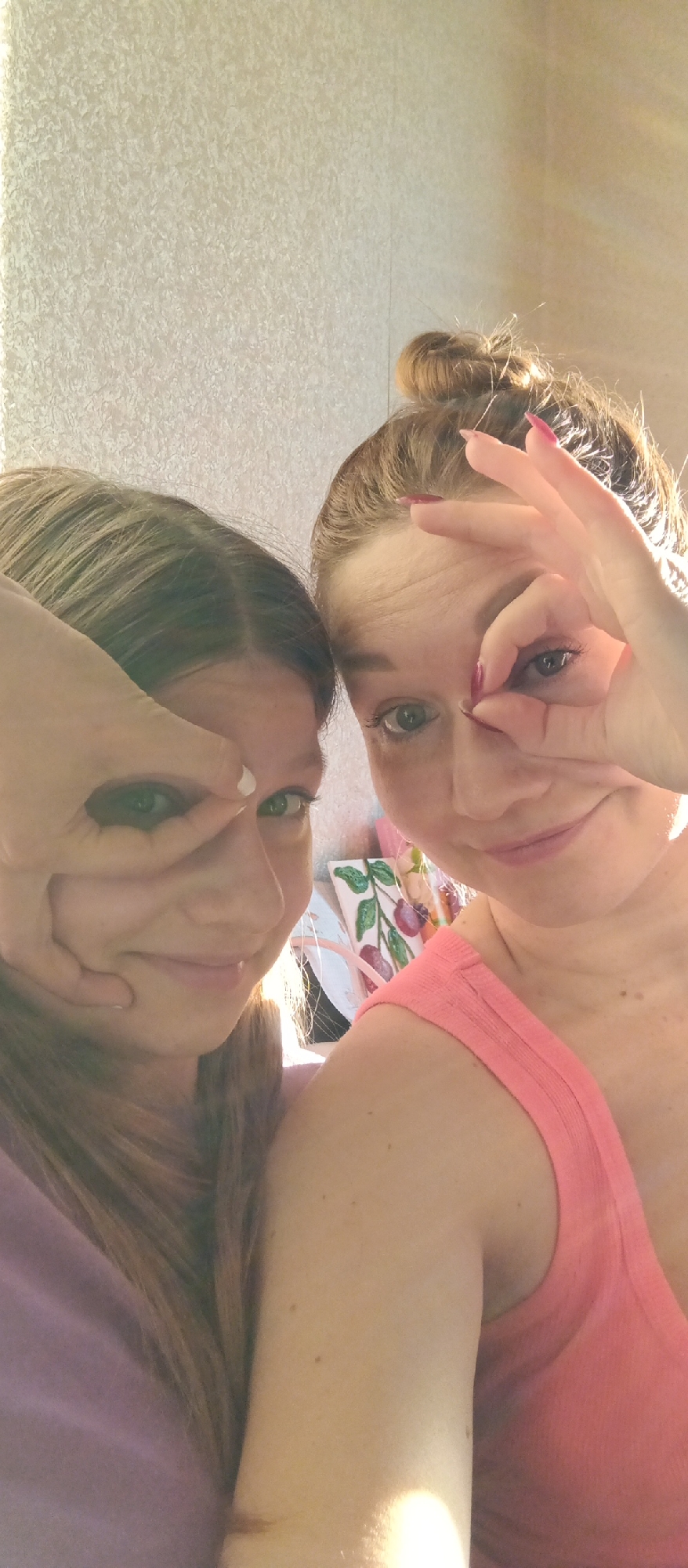 Two women are posing close together for a selfie indoors. Both are making the "circle game" gesture with their hands, framing one eye each. The woman on the left has long blonde hair, and she is wearing a lilac-colored shirt. The woman on the right has her auburn hair up in a bun and is wearing a pink tank top. In the background, there is a textured wall and a painting with floral elements is slightly visible. Location: Unknown, Unknown.