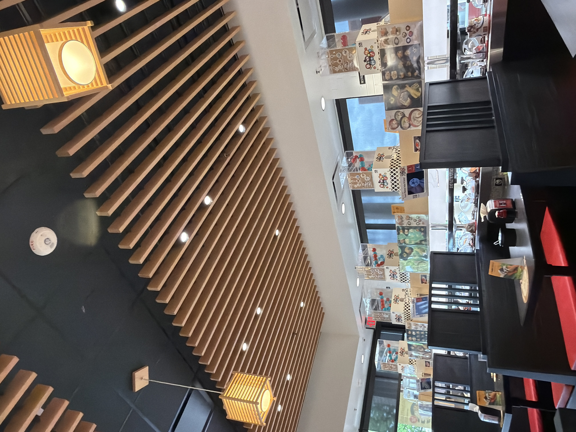The image shows the interior of a restaurant in Beaverton, Oregon. The restaurant features a modern design with a black ceiling and a slatted wooden ceiling feature. Several square, wooden pendant lights hang from the ceiling. In the foreground, there are black booths with red seats, and black tables are visible. On the tables, there are menus and condiments. In the background, there is a conveyor belt system, typical of a sushi restaurant, with clear domes covering dishes. Various boxes and displays are stacked behind the conveyor belt, some containing what appear to be small toys or trinkets, and others displaying images of food. A white fire alarm is mounted on the black ceiling.
