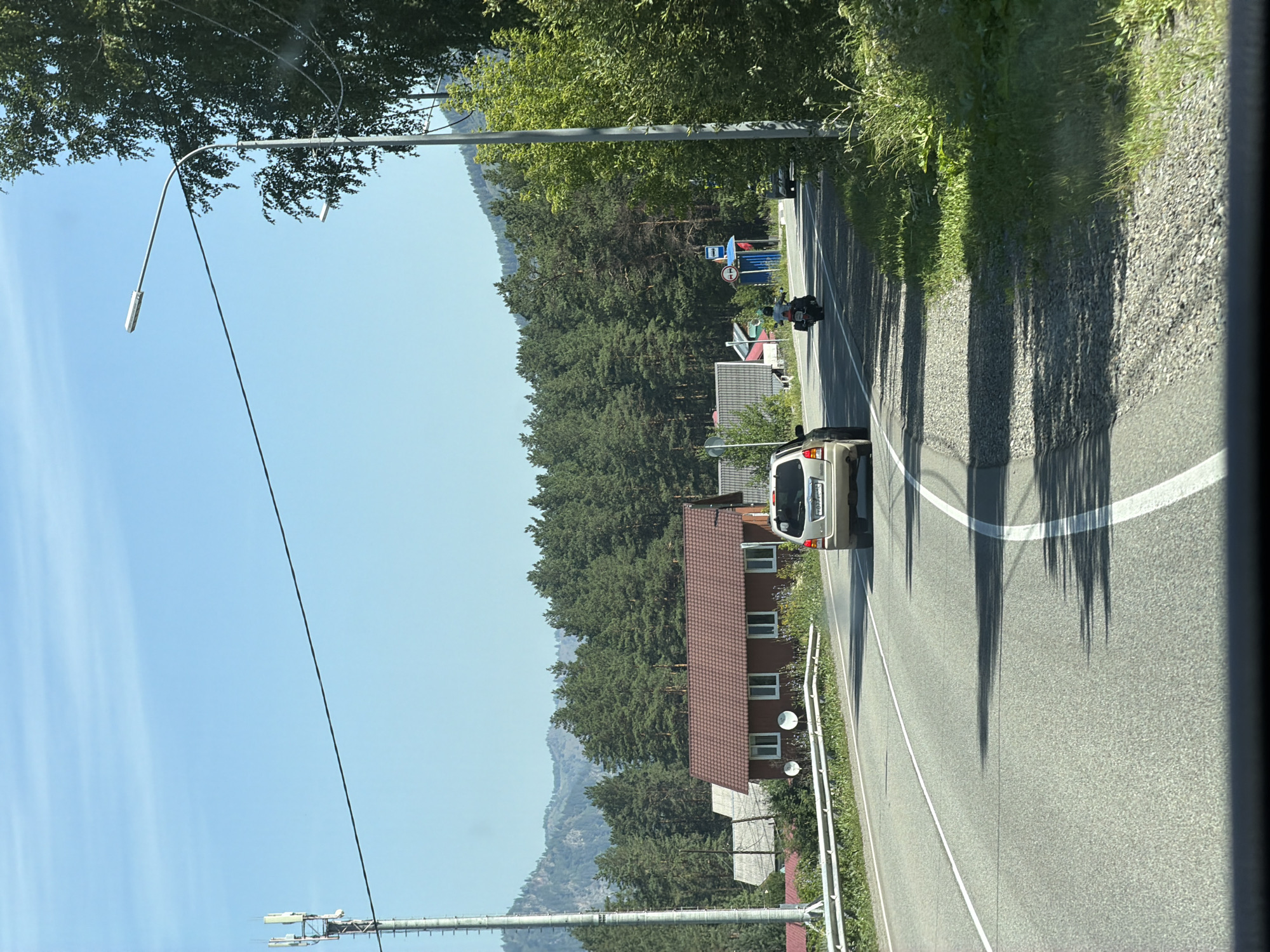 The image depicts a bright, sunny day in Ust-Muny, Russia, along a paved road. A light-colored station wagon is in the foreground, driving on the right side of the road. To the left of the road, a red house with a brown, tiled roof and white-trimmed windows is visible. Further to the left, a tall cell tower stands against a clear blue sky. Beyond the house and the cell tower, a dense forest of green pine trees stretches across a mountainous backdrop. A motorcycle is also present on the road, following the station wagon. In the distance, a blue bus stop shelter with a red roof is situated on the side of the road. The road itself has clear white lane markings. Lush green vegetation and trees line the right side of the road.