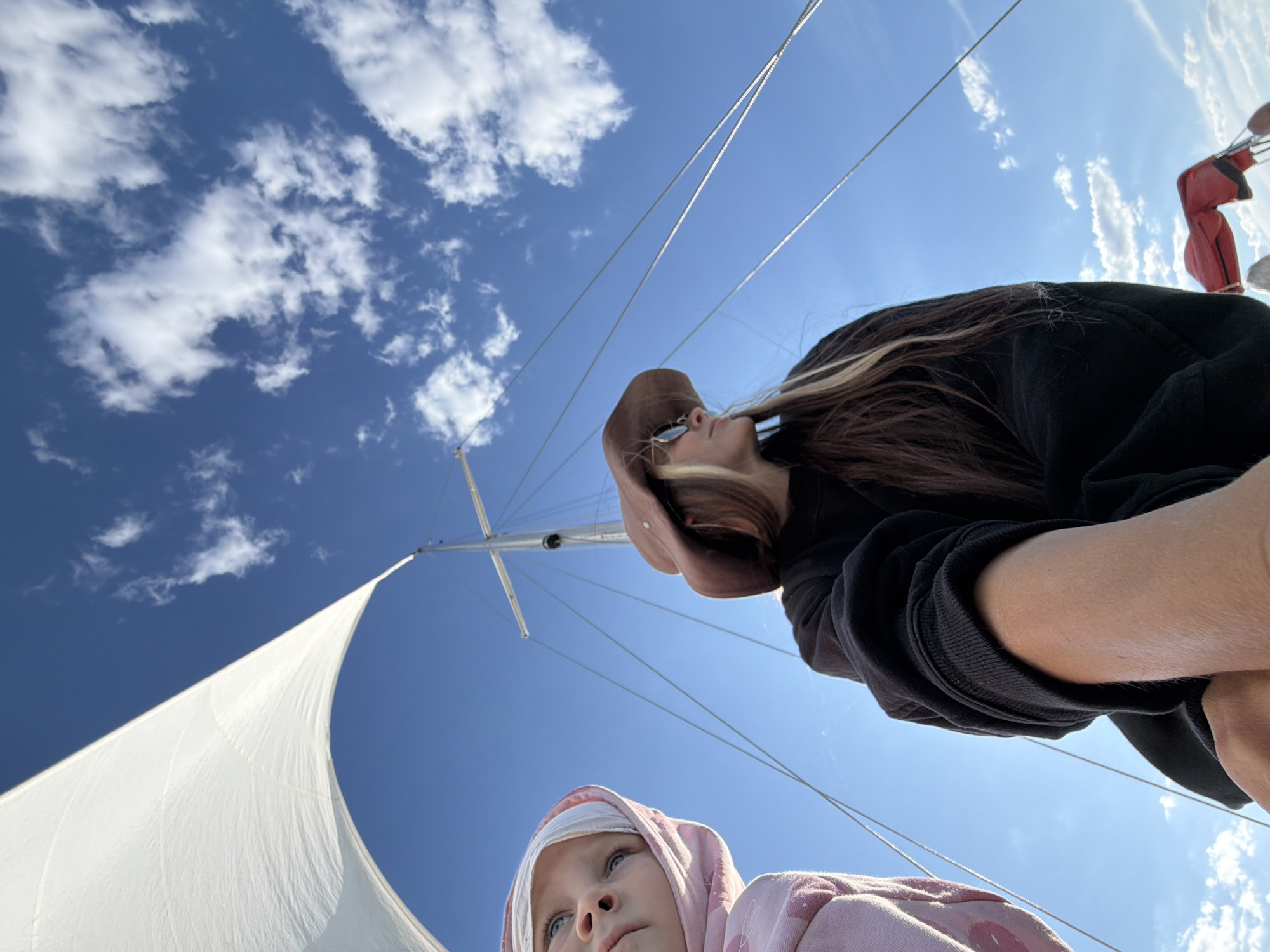 The image depicts two individuals on a sailboat against a clear blue sky with scattered white clouds. A large white sail dominates the upper left portion of the frame, with rigging visible against the sky. In the foreground, a person wearing a brown hat and sunglasses is looking to the right. To their left, a young child with blue eyes is looking upwards. The child is wearing a pink hooded garment. The overall scene suggests an outdoor activity, possibly a leisurely trip on the water. The specific location in Kamyshin, Russia, cannot be determined from the visual information alone.