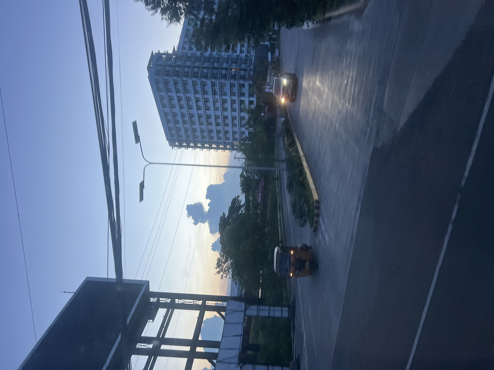 The image captures a street scene in Davao City, Philippines, likely during twilight given the soft lighting and blue hues of the sky. A multi-story building, possibly an apartment complex or hotel, dominates the right side of the frame, its numerous windows reflecting the dim light. In the foreground, the asphalt road shows reflections from vehicle headlights.
On the road, a three-wheeled motorized vehicle, commonly known as a "tricycle" in the Philippines, is moving away from the viewer. Further down the road, a dark-colored SUV with its headlights on is approaching. The left side of the image features a large, dark structure, possibly a billboard or a part of an industrial facility, with supporting metal beams and white panels. Beyond the road, lush green trees and vegetation are visible, with some peculiar, cloud-like formations in the sky that bear a resemblance to figures. Overhead, a network of thick power lines crisscrosses the sky. The overall atmosphere is one of a quiet urban evening, with vehicles and infrastructure characteristic of a developing city.