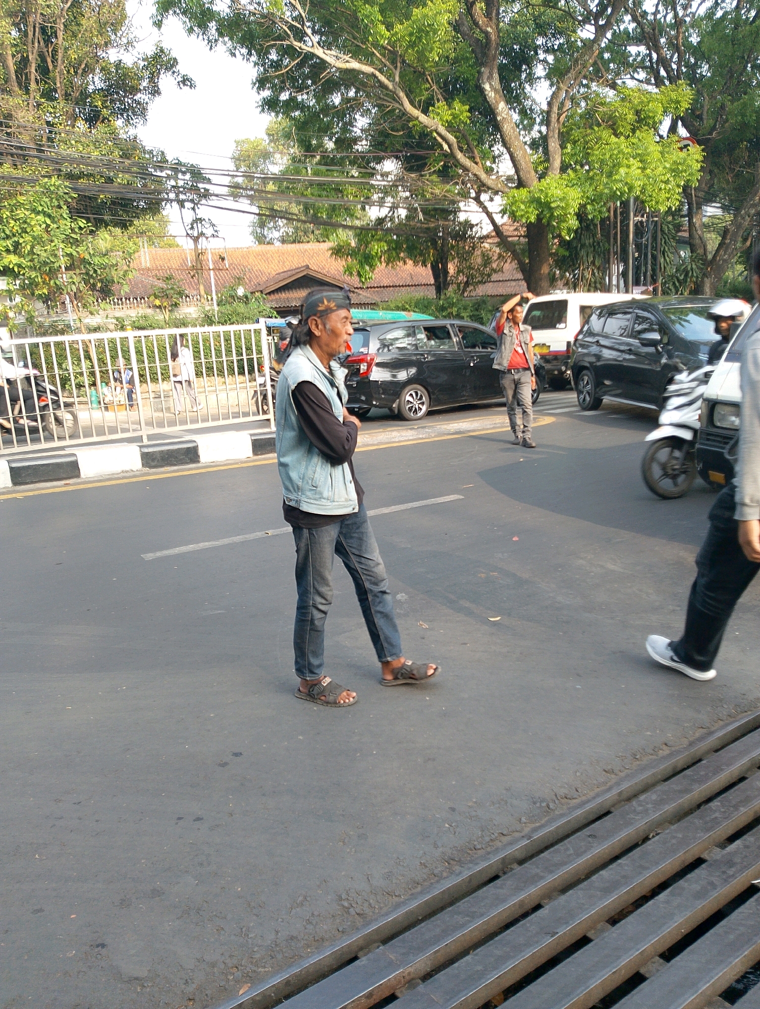 In Bandung City, Indonesia, a middle-aged man with dark, long hair and a black cap is standing on the asphalt road. He is wearing a light blue denim vest over a black long-sleeved shirt and dark blue jeans. His right hand is raised to his forehead as if shielding his eyes from the sun, and his left arm is bent with his hand resting on his chest. He is wearing dark sandals on his feet.
The road appears to be a busy street with several vehicles present. A white car and a black SUV are parked or stopped in the background, along with a white motorcycle. To the left of the man, a white metal fence runs along the side of the road, behind which a building with a brown tiled roof is visible. Trees with green foliage line the street, and power lines are strung overhead. The scene suggests a warm, sunny day in an urban environment.