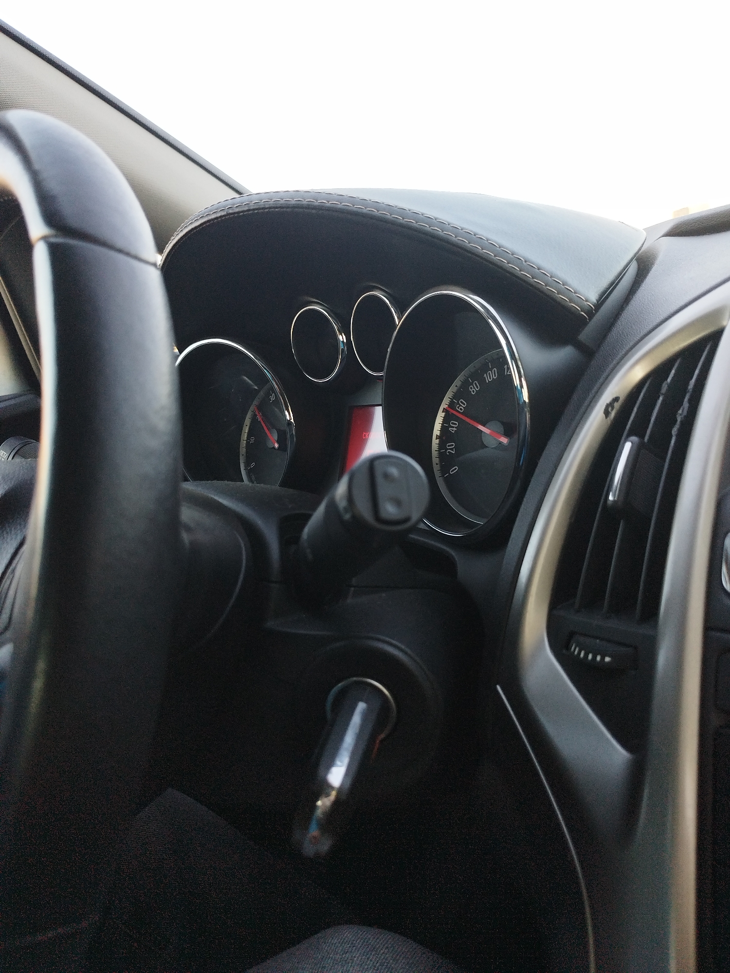 This is a close-up, eye-level, interior shot of a car dashboard. The primary focus is on the steering wheel, which is on the left and is black with a leather-like texture. To the right of the steering wheel, a portion of the car's instrument cluster is visible, featuring a speedometer with a red needle pointing between 20 and 40. There are also two smaller gauges to the left of the speedometer, each with a silver ring. A silver trim piece surrounds the instrument cluster. Further to the right, a silver-trimmed air vent is visible, with horizontal slats and a silver lever. The dashboard itself is covered in black leather with visible stitching along the top edge. A car key is inserted into the ignition, located below the steering column. The background is mostly out of focus, showing a bright, possibly overcast, sky. The overall lighting is natural, suggesting it is daytime. There are no people visible in the image. The car appears to be parked.