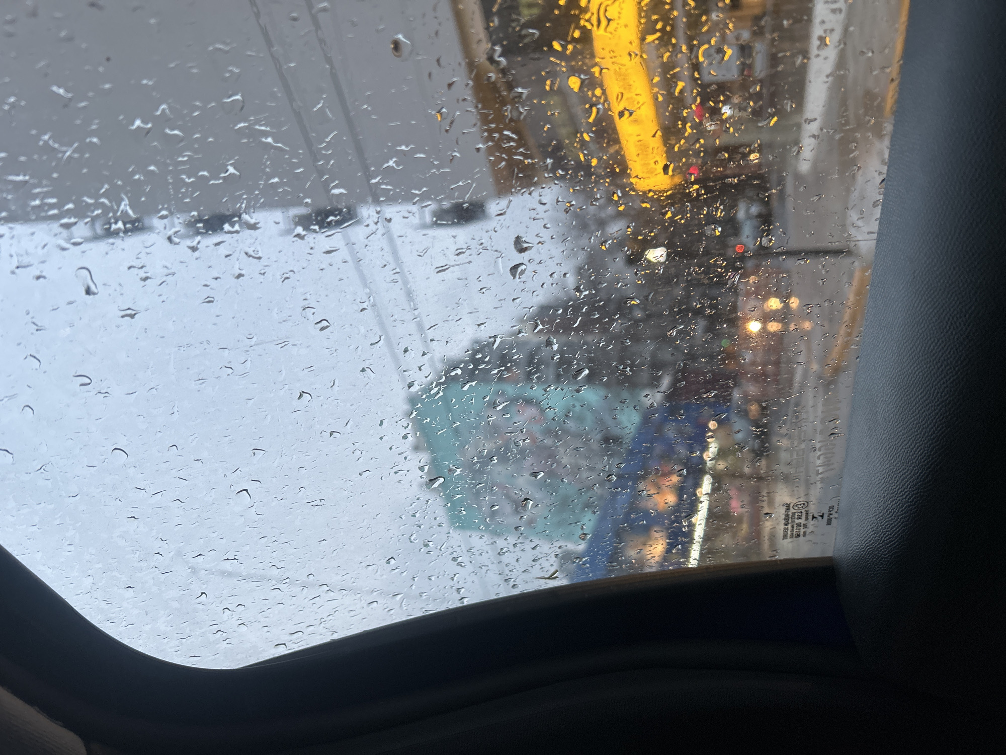The image shows a view from inside a vehicle looking out through a rain-streaked window. The foreground is dominated by the dark texture of the car's interior paneling and window frame. The glass is covered with numerous water droplets of varying sizes, distorting the view of the outside scene.
Through the rain, a city street is visible. On the left, a large building is partially obscured by the rain and the window frame. It appears to have a mural on its side, with hints of turquoise and other colors. Below this building, there are lights, possibly from shops or streetlights, reflecting on the wet street.
To the right, a bright yellow awning or sign is prominent, likely from a commercial establishment. Other lights, appearing as blurred yellow and red orbs, suggest traffic and illuminated buildings further down the street. The overall atmosphere is that of a rainy day in an urban environment. Without specific landmarks or identifying signs within the visible area, pinpointing the exact location in Buenos Aires, Argentina is not possible from this image alone.