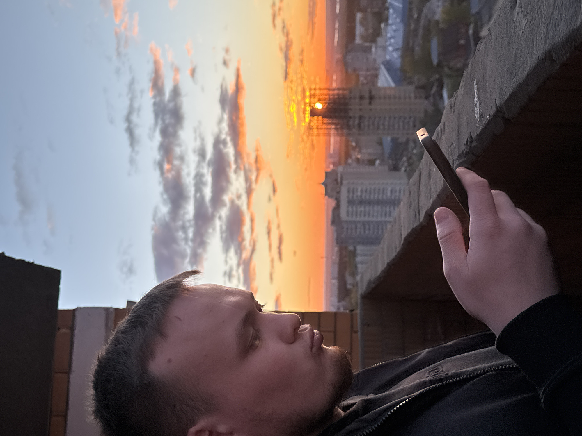 A person with short hair and a beard is looking at a smartphone, held in their right hand, with the sunset reflecting on the screen. They are on a roof ledge overlooking a city, likely Perm, Russia, based on the architectural style of the high-rise buildings in the background. The sky transitions from blue to orange as the sun sets behind the buildings. The person is wearing a black jacket. The rooftop ledge appears to be constructed of brick and concrete.