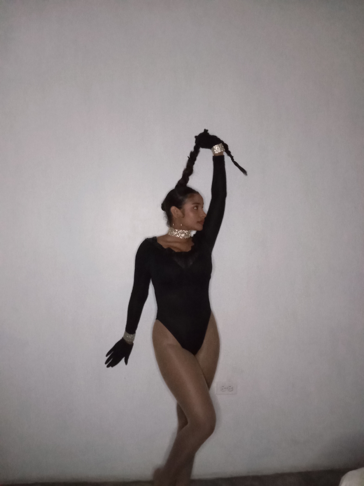 An individual, identified as a woman, is captured posing indoors against a plain, light-colored wall in Juan Griego, Venezuela. She is dressed in a black long-sleeved bodysuit, possibly with sheer or nude-colored hosiery, and black gloves. Her ensemble is accessorized with a prominent silver-toned choker necklace and matching bracelets on both wrists. Her dark hair is styled into two long braids, one of which she holds aloft with her right hand, creating an upward-reaching gesture. Her left arm is extended downwards. She is positioned facing slightly to the side, looking towards the right of the frame. The lighting is dim, casting a somewhat moody atmosphere. A white electrical outlet is visible on the wall in the lower right portion of the image. The floor beneath her appears dark. The pose suggests a stylized performance or portrait.