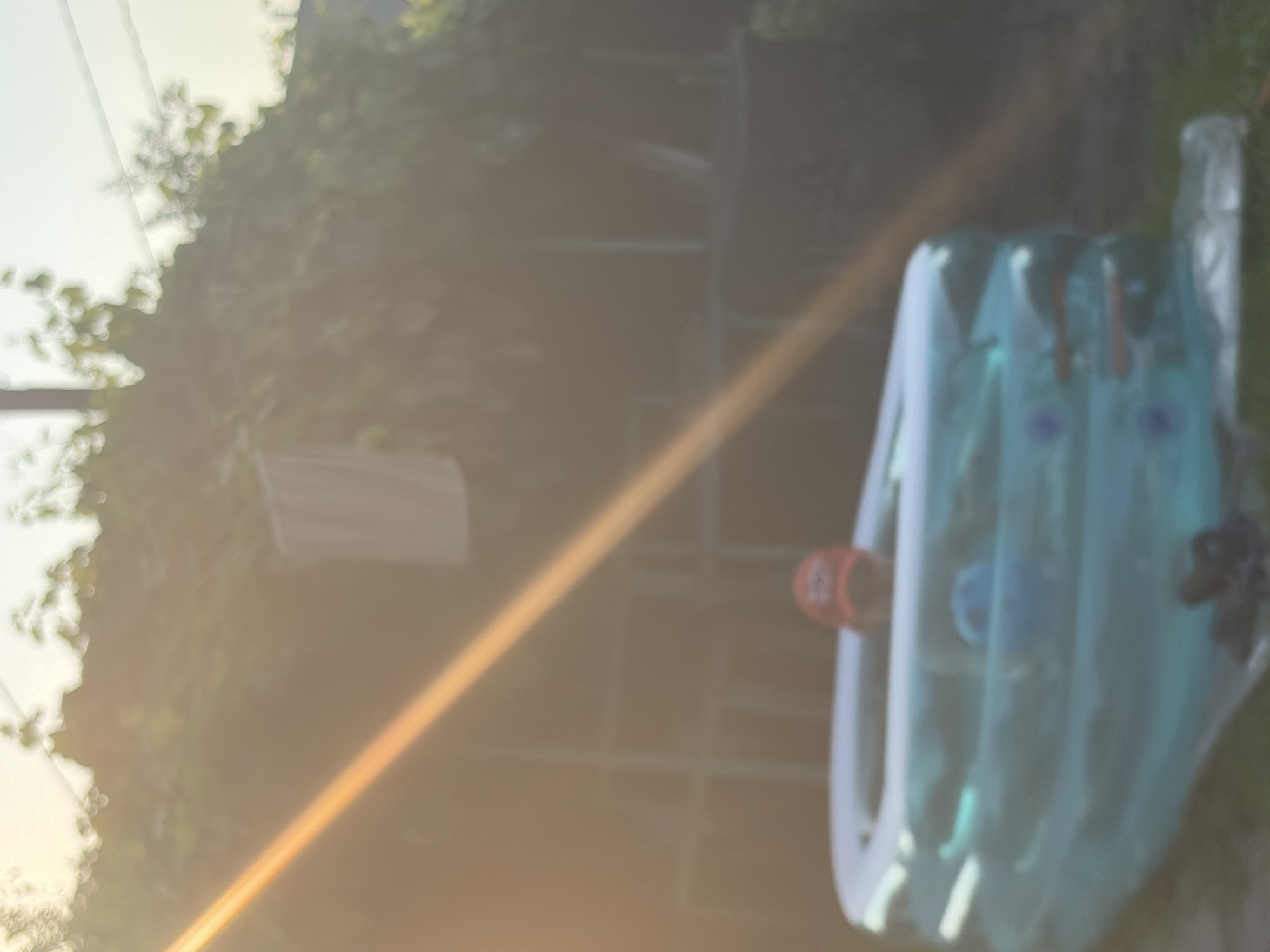An unfocused shot shows a person wearing a red baseball cap in a blue inflatable swimming pool outdoors. The pool is the main subject and is filled with water. Sunlight creates a glare across the image, and foliage is visible in the background.
