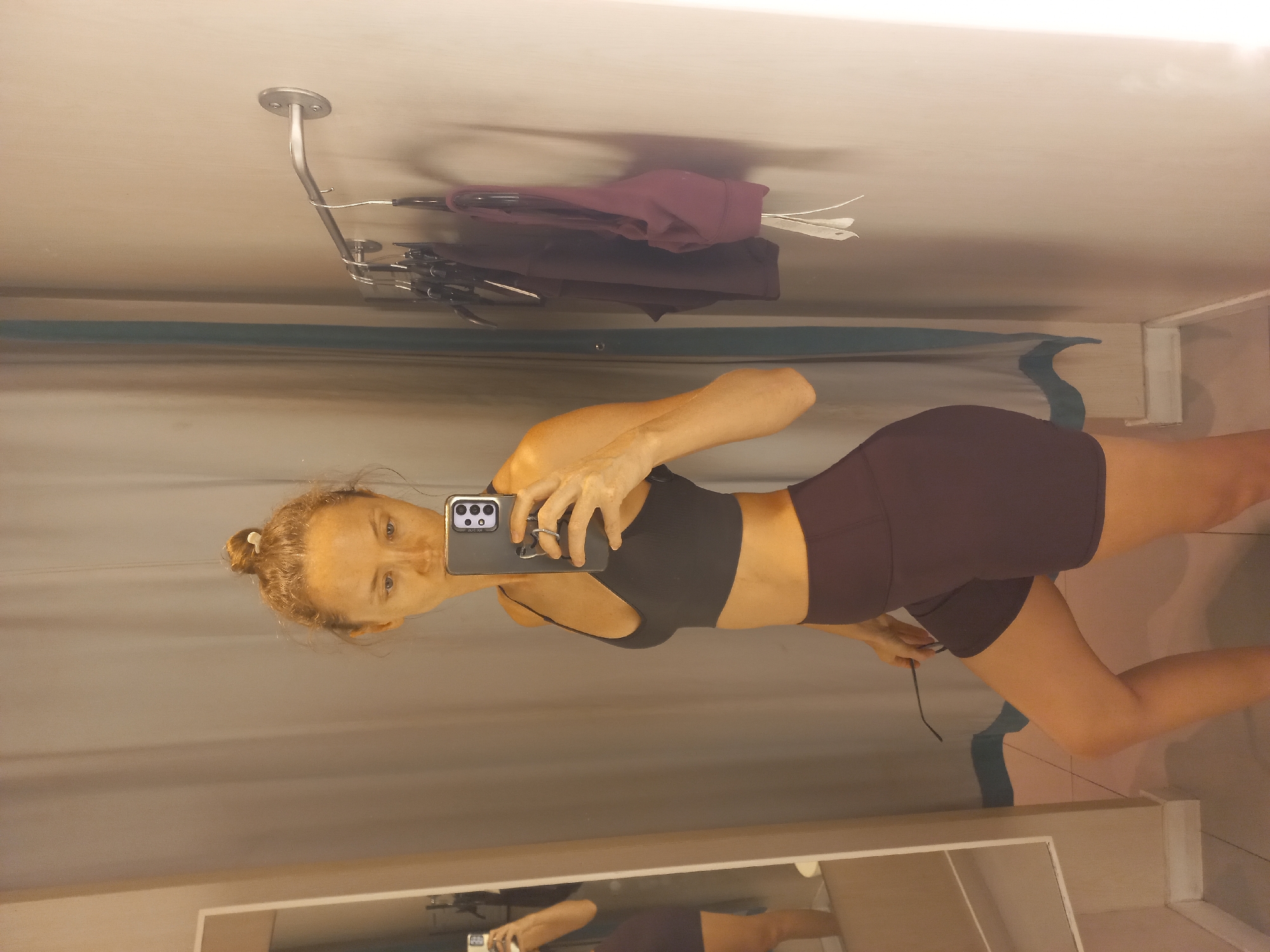 A woman with blonde hair in a bun takes a selfie in a dressing room. She wears a black sports bra and maroon shorts. Clothes hang on a rack above. The location is likely a retail store in Pattaya City, Thailand, based on the presence of dressing rooms, clothing, and likely tourist activity.