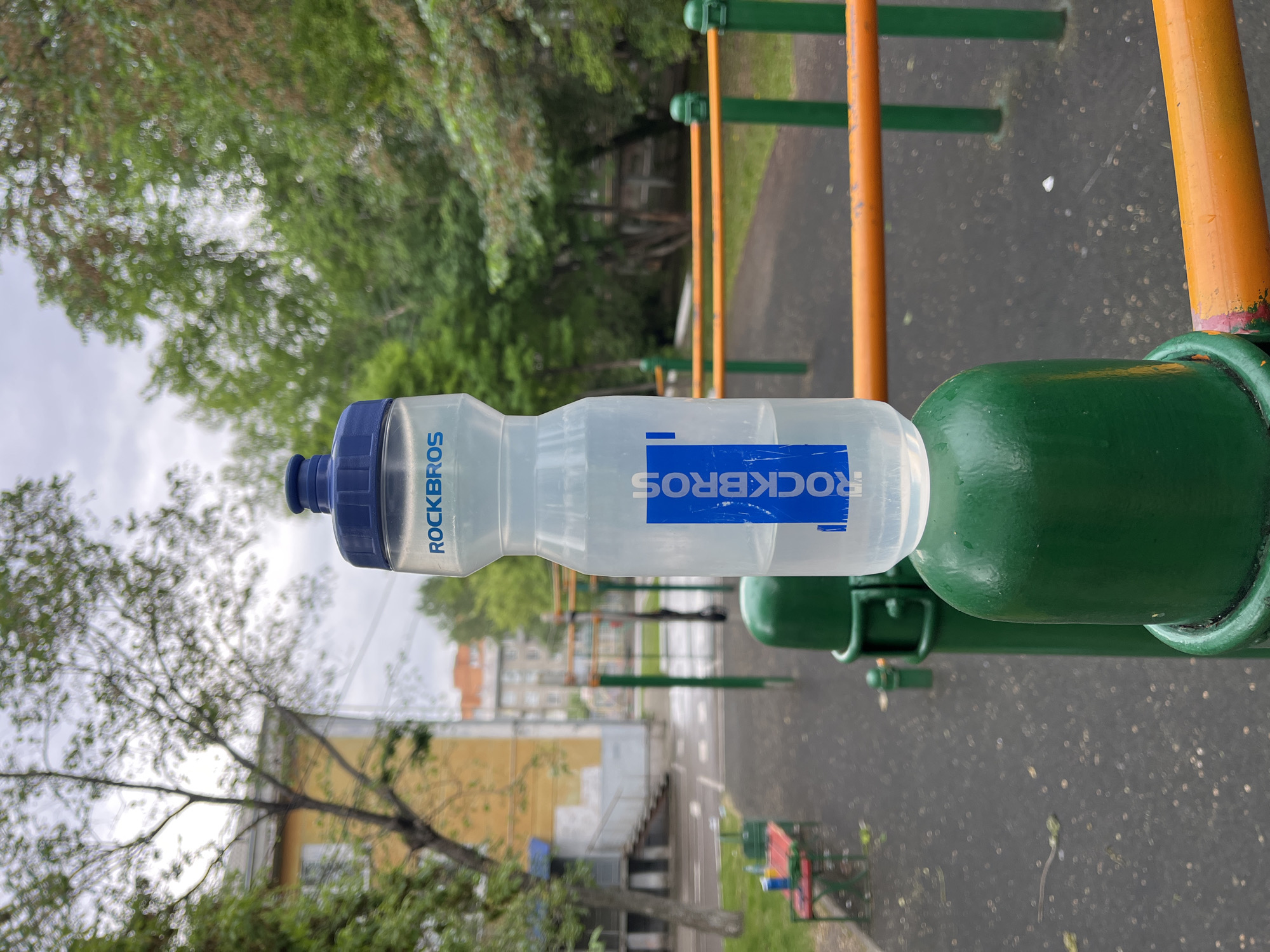A clear plastic water bottle with a blue lid and a blue label featuring the brand name "ROCKBROS" is positioned atop a green metal pole. The bottle contains a clear liquid, approximately half full. The background depicts an outdoor urban setting, possibly a children's playground or an outdoor gym, with green metal bars and orange horizontal bars visible. Trees and residential buildings are present in the distance. The ground appears to be paved with dark material, possibly rubberized safety surfacing common in playgrounds. The scene suggests a public outdoor recreational area in the city of Krasnoyarsk, Russia, likely during daylight hours with overcast sky. There are no people visible in the image.