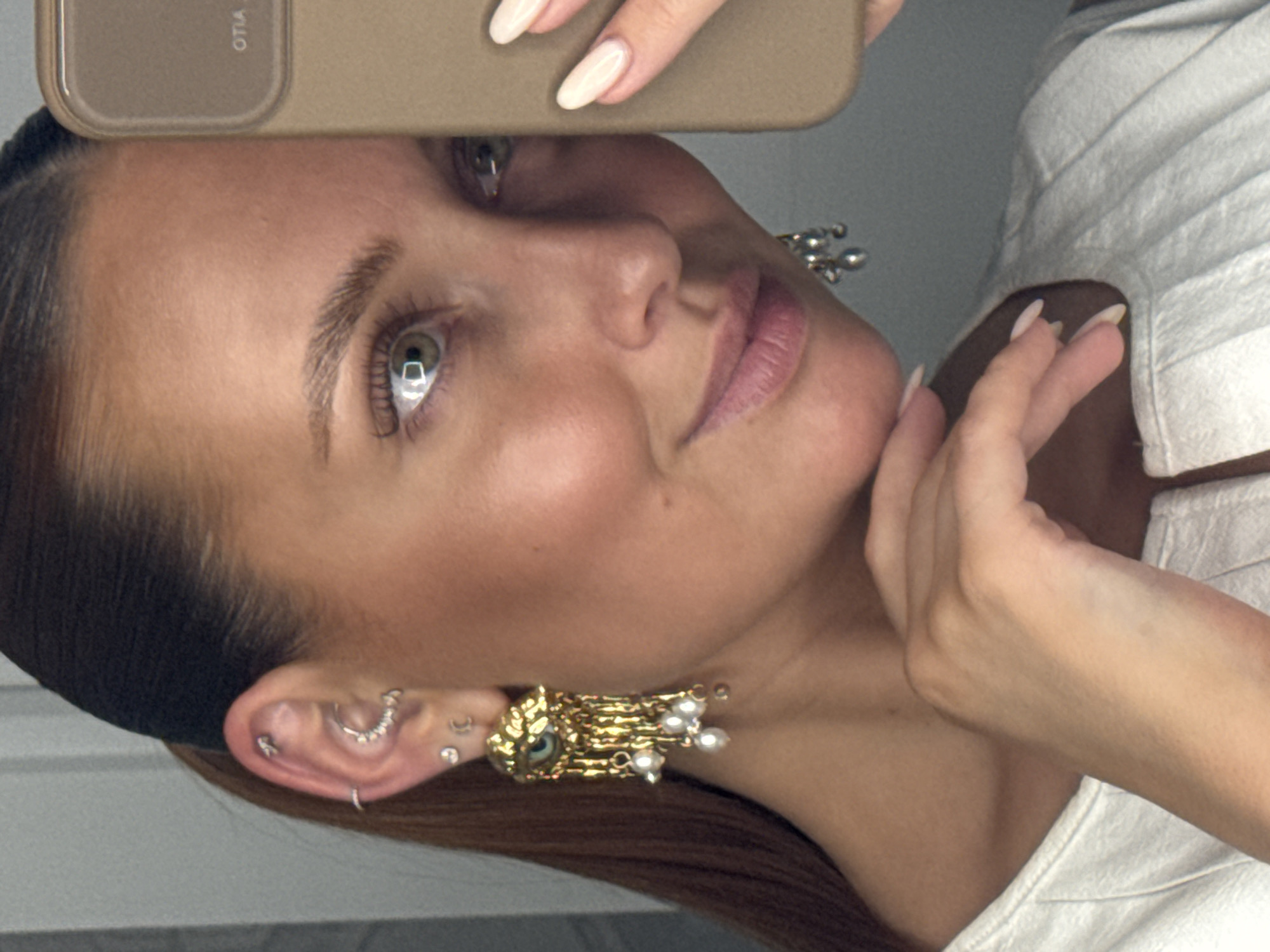 A fair-skinned person is holding up a phone with a beige case. Part of their face is visible, including an eye, eyebrow, nose, cheek, and mouth. The person has light brown hair pulled back, with multiple ear piercings. The person is wearing a gold earring with an eye design and small pearls. The person is wearing a white shirt and holding their hand near their neck. There is no indication of the location being Chaikovsky, Russia.