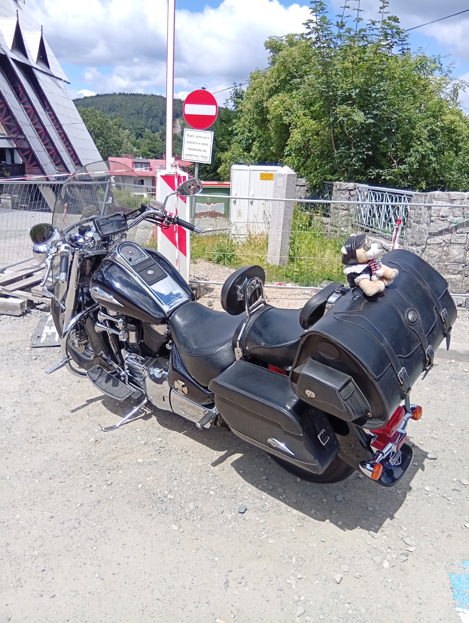 A black motorcycle is parked on a gravel lot in front of a modern building with a distinctive triangular roofline. The motorcycle has large black saddlebags and a stuffed teddy bear wearing a pilot's hat and scarf is perched on the rear saddlebag. A red and white striped pole with a circular prohibition sign and a sign with Polish text is positioned to the left of the motorcycle. In the background, there are trees and a hilly landscape. The scene appears to be in the city of Karpacz, Poland, likely near a tourist attraction.