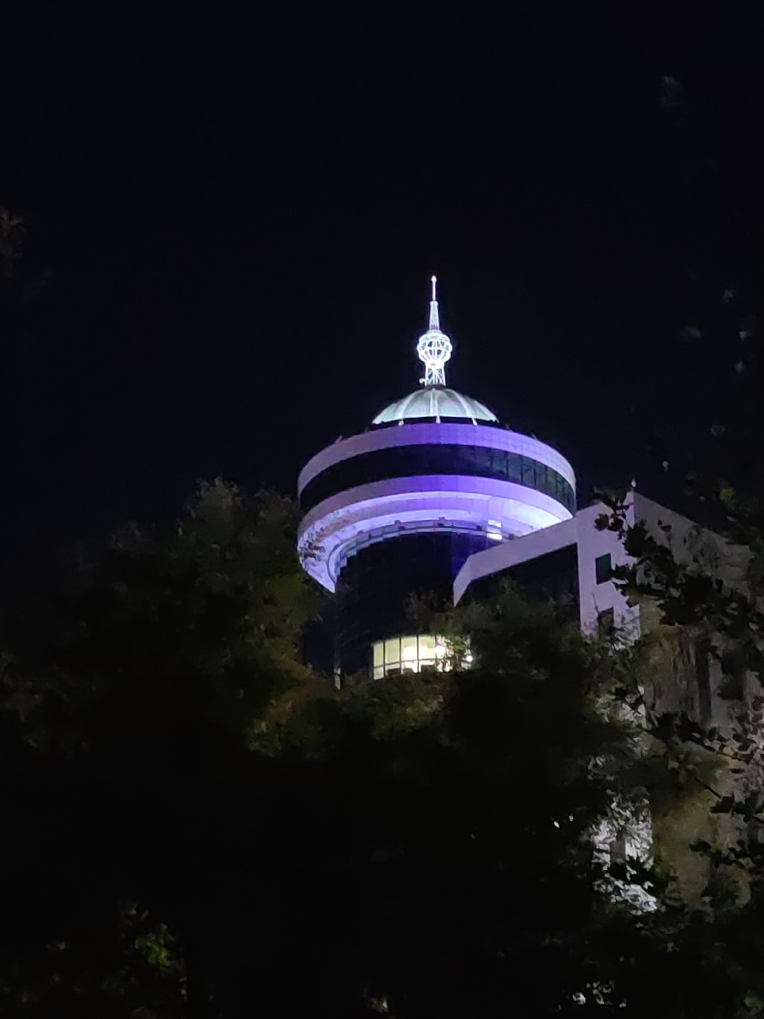 The image shows the Tashkent Television Tower at night, a prominent landmark in Tashkent, Uzbekistan. The tower's upper section is illuminated with purple and white lights, highlighting its distinctive dome and spire. The surrounding area is dark, with the silhouettes of trees obscuring parts of the lower structure. A portion of an adjacent building, also lit, is visible to the right of the tower.