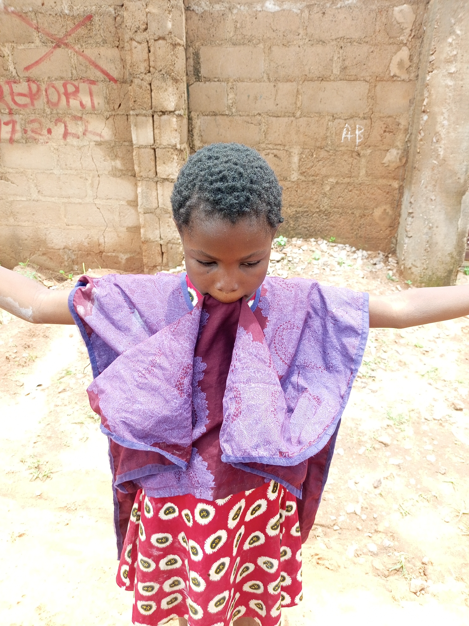 A medium shot captures a young Black girl in Tempure, Nigeria, standing outdoors in front of a rough, tan brick wall. She is wearing a purple and maroon patterned dress, the top layer a wide, draped purple fabric with an intricate pattern, and the bottom layer a vibrant red skirt with a white and yellow oval-shaped pattern. Her arms are outstretched to her sides. The background wall has red graffiti that reads "REPORT" and what appears to be a date, "17.3.21". To the right of the graffiti, "AB" is scrawled in white. The ground beneath her is composed of dirt and scattered small stones.