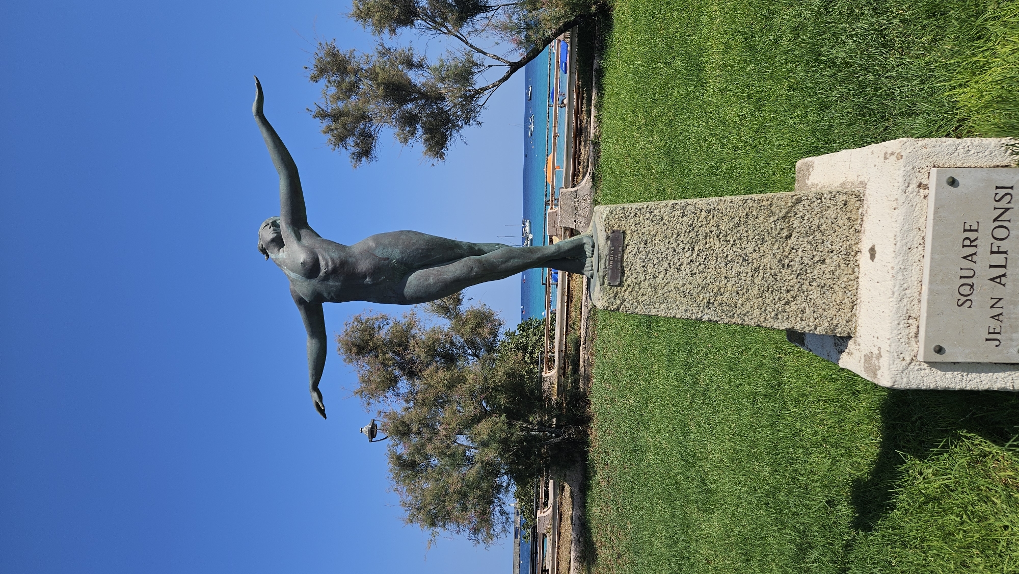 The image shows a bronze statue of a nude female figure with arms outstretched, set on a granite pedestal. The statue is located in Square Jean Alfonsi in L'Île-Rousse, France, with the Mediterranean Sea and several sailboats visible in the background. The scene is set against a clear blue sky on a sunny day, with manicured green grass in the foreground and trees on either side of the statue. A plaque at the base of the pedestal reads "SQUARE JEAN ALFONSI". Another plaque on the statue's pedestal reads "ONDINE par JUAN BORREL".