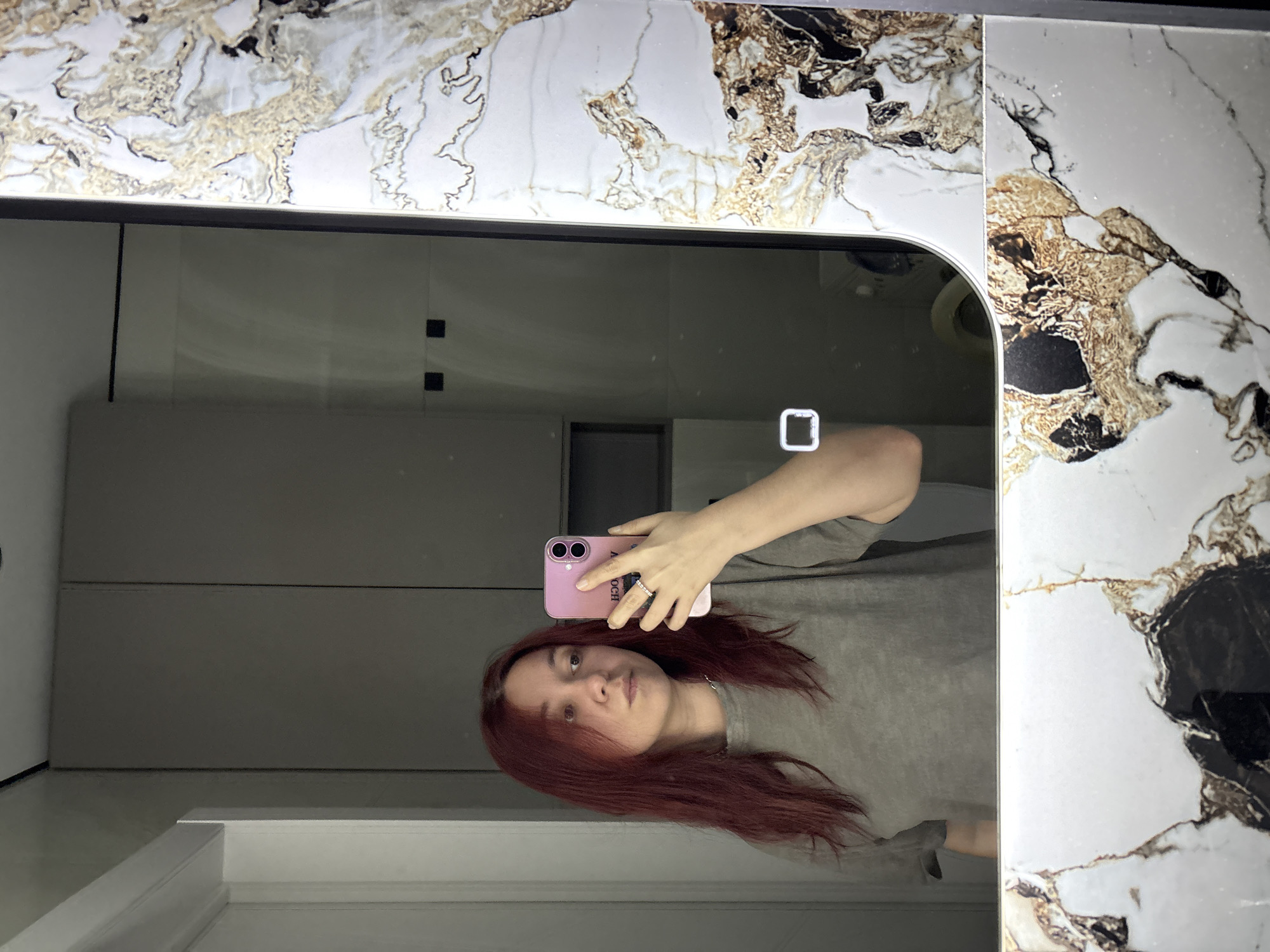A female individual with medium-length, reddish-brown hair is capturing a mirror selfie in an interior space located in Kazan, Russia. She is wearing a plain grey t-shirt with a rounded neckline and a thin silver-colored chain around her neck. A silver-colored ring with small, clear stones is visible on her right ring finger. She holds a pink smartphone with dual rear cameras in her right hand, positioning it to reflect her image. Her gaze is directed slightly to her right, away from the phone's lens.
The mirror features a thick, ornate frame composed of a white base with prominent golden-brown and black veining, mimicking marble. A white square icon with rounded corners, likely a touch control for the mirror's functionality (e.g., lighting), is illuminated in the lower right quadrant of the reflective surface.
The reflected background reveals a modern, minimalist interior. On the far left of the reflection, a white door frame is partially visible. The central background consists of light grey panels or cabinetry. Higher on the right side of the reflection, two small black square elements are present on a wall. Below them, a dark, rectangular recess or shelf is visible. In the lower right of the reflection, part of a white, curved appliance, potentially a washing machine or toilet, can be discerned. The scene is illuminated by interior artificial lighting.