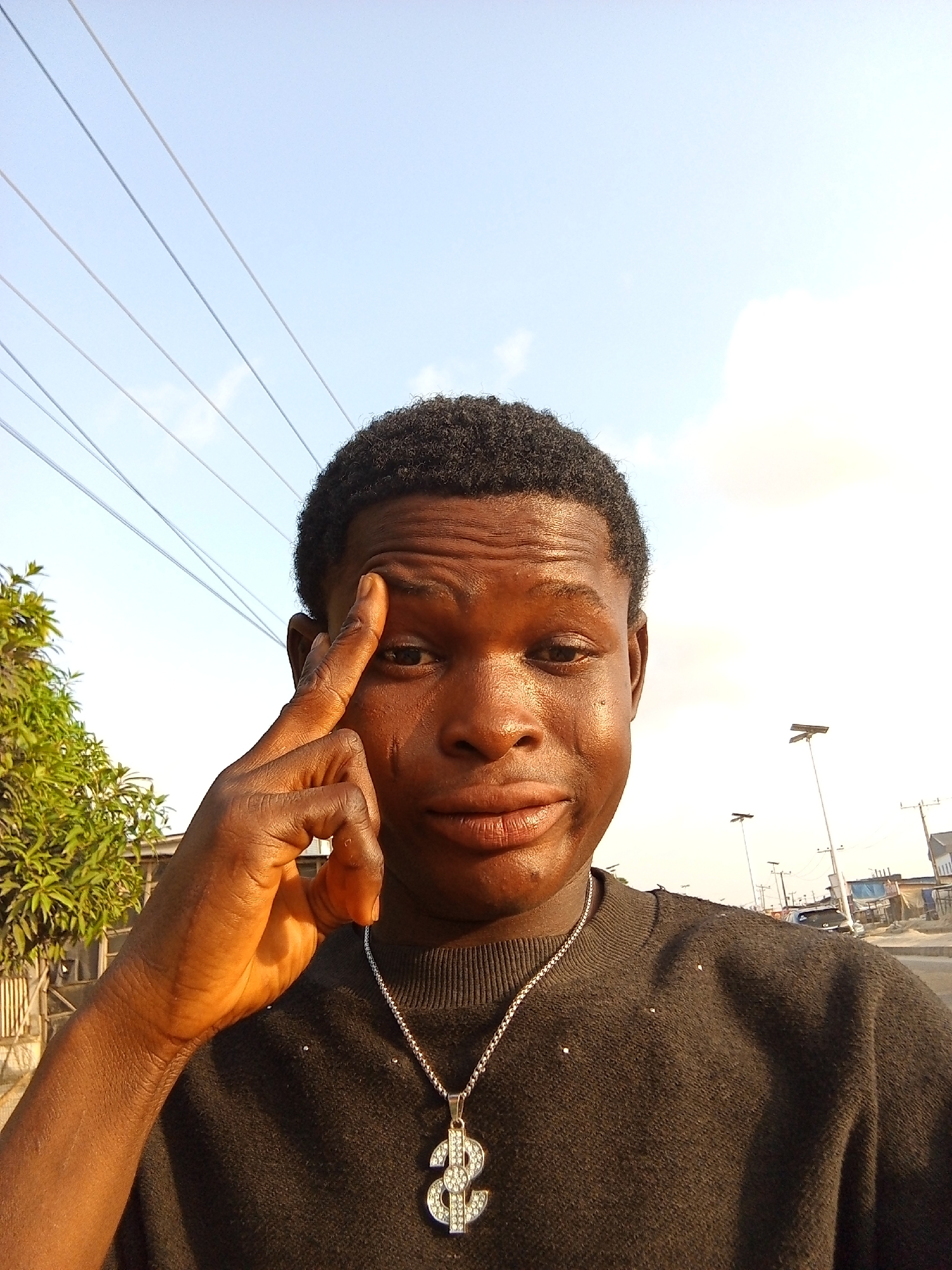 A young man is taking a selfie outdoors, likely in the city of Ado, Nigeria. He has dark skin, short black hair, and wears a black sweater with a silver chain featuring a dollar sign pendant. He is holding his finger to his forehead in a thoughtful gesture. Overhead power lines are visible in the background, along with the sky. A tree is located on the left of the frame. Street lights and buildings can be seen in the distance, suggesting an urban environment.