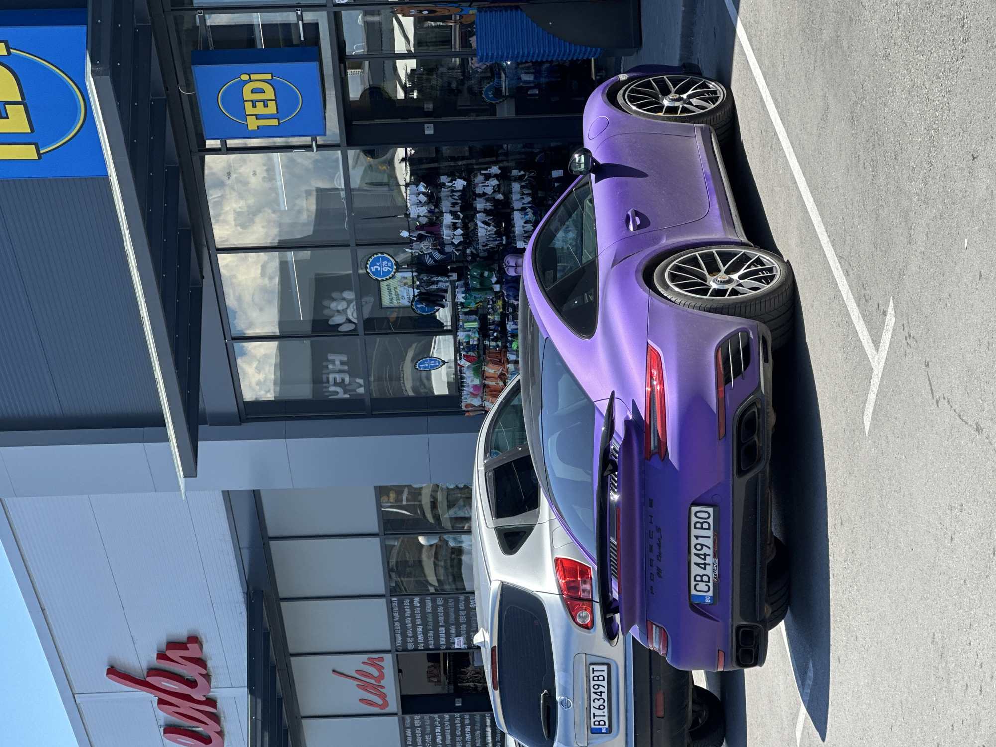 This image depicts a parking lot scene in front of a retail establishment in Veliko Tarnovo, Bulgaria. The most prominent subject is a matte purple Porsche 911 Turbo S sports car parked in a designated spot. To the left of the Porsche, a silver Nissan Qashqai SUV is also parked.
The background features the facade of two adjacent retail stores. The store on the left has signage with the name "eden" in red script. The store on the right displays the "TEDI" logo in blue and yellow on a prominent sign. The windows of the TEDI store reveal racks of merchandise, suggesting it is a discount or variety store. The environment appears to be a sunny day, with clear blue skies visible and reflections of clouds in the store windows. The parking lot is paved, and white parking lines are clearly visible. No people are visible in the image. The license plate of the Porsche is visible and reads "CB 4491 BO". The license plate of the Nissan reads "BT 6349 BT".
