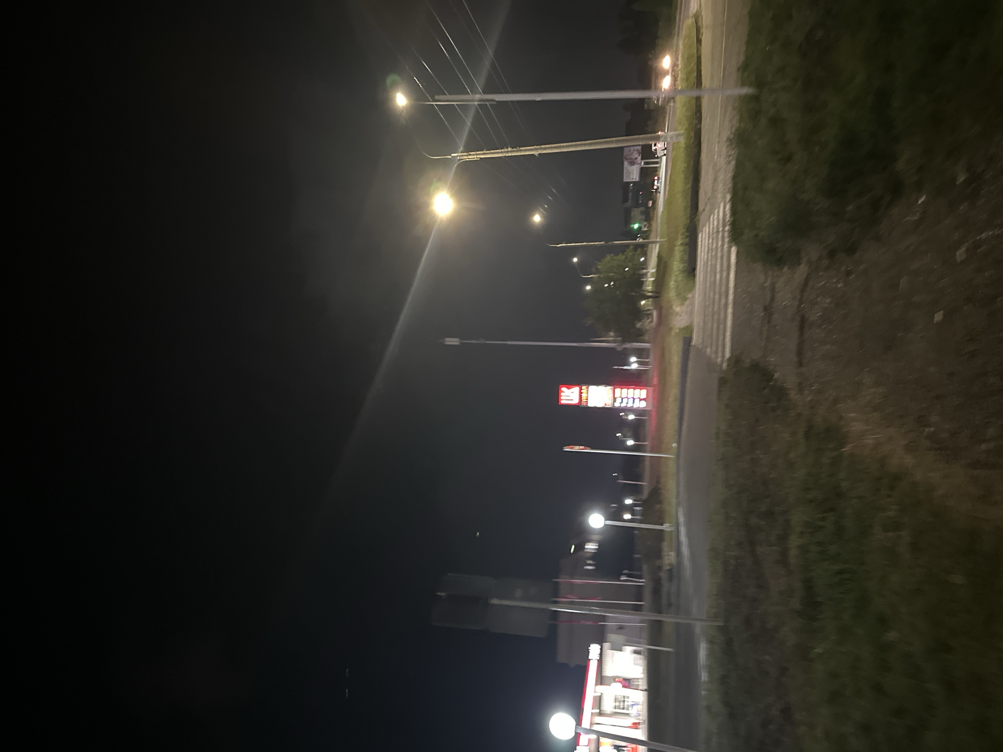 At night, in the city of Nevinnomyssk, Russia, a gas station sign with "UK" in red letters and Russian text underneath is visible. The gas station has illuminated pillars and a white building. Streetlights illuminate the road and sidewalk. A pedestrian crossing is visible in the foreground. Cars are driving on the road in the distance. The sky is dark and clear.