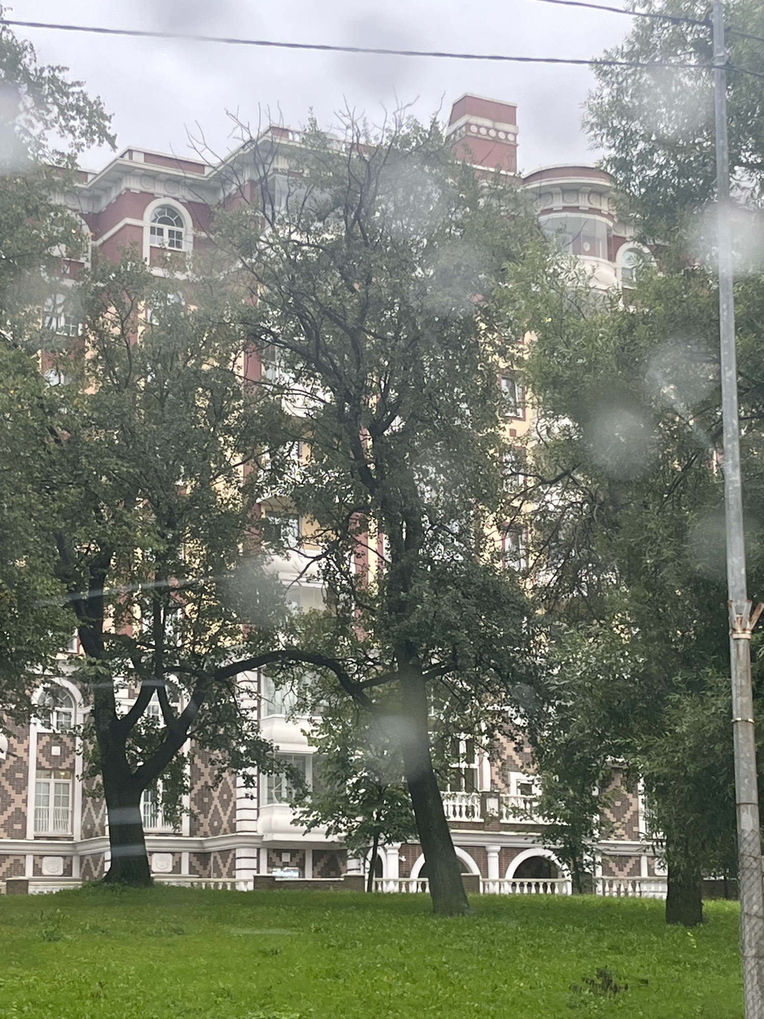The image shows an ornate red brick building with white architectural details, possibly in Saint Petersburg, Russia, due to its style. The building has arched windows and balconies, and intricate decorative patterns on its facade. Large, leafy trees are in the foreground, partially obscuring the view of the building. A patch of green grass is visible at the bottom of the image. The sky is overcast, suggesting a cloudy day. There are no people visible in the scene. The overall impression is one of an older, grand building set within a green, possibly park-like, environment.