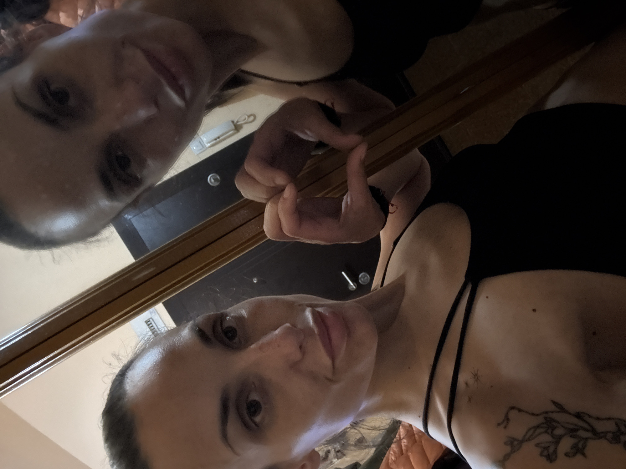 A woman takes a selfie in front of a mirror. She is wearing a black top with thin straps and has a tattoo on her right shoulder. Her reflection is visible in the mirror, and she is making a heart shape with her hands. The background appears to be a room interior, possibly a bedroom, with a piece of furniture visible behind her. It is not possible to confirm whether the location is in Pleven, Bulgaria, based solely on the visual information.