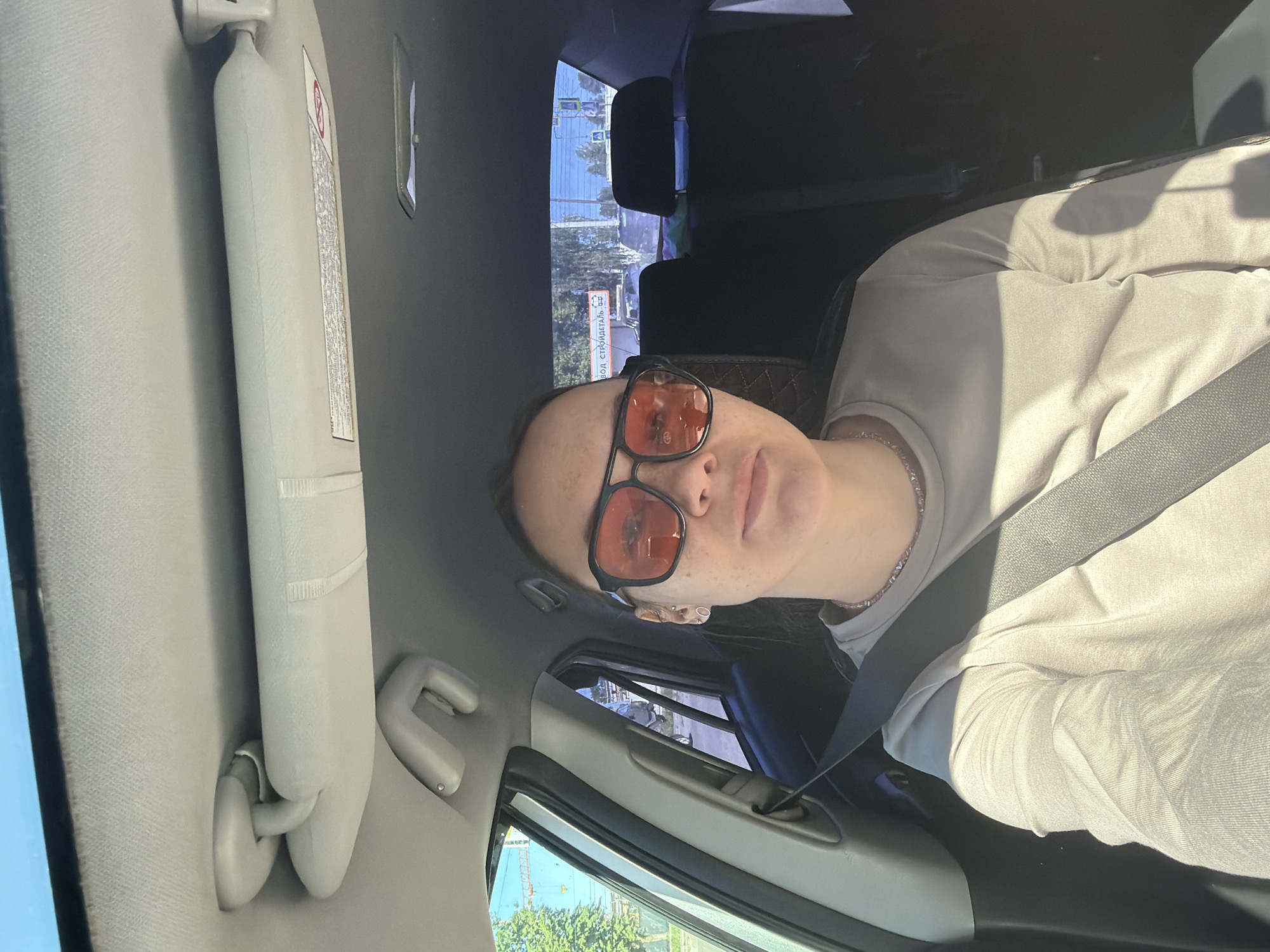 A young woman is in the passenger seat of a car, taking a selfie. She is wearing oversized sunglasses with orange lenses and a light-colored long-sleeved shirt. Her seatbelt is fastened. Outside the car window, trees and buildings are visible, indicating an urban environment. The interior of the car includes a sun visor and a portion of the dashboard. The scene appears to be in the city of Chaikovsky, Russia, given the signage in the background.
