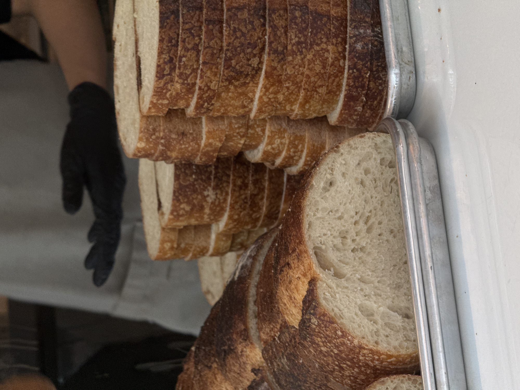 In a bakery, possibly in Bangkok, Thailand, slices of bread are stacked in metal trays. In the background, a person wearing black gloves and a light-colored apron is visible, suggesting preparation or service of the bread. The bread appears to be artisan loaves, with crusty exteriors and soft interiors, some of which are already sliced and arranged in neat stacks. The overall scene conveys a sense of fresh baked goods being presented.